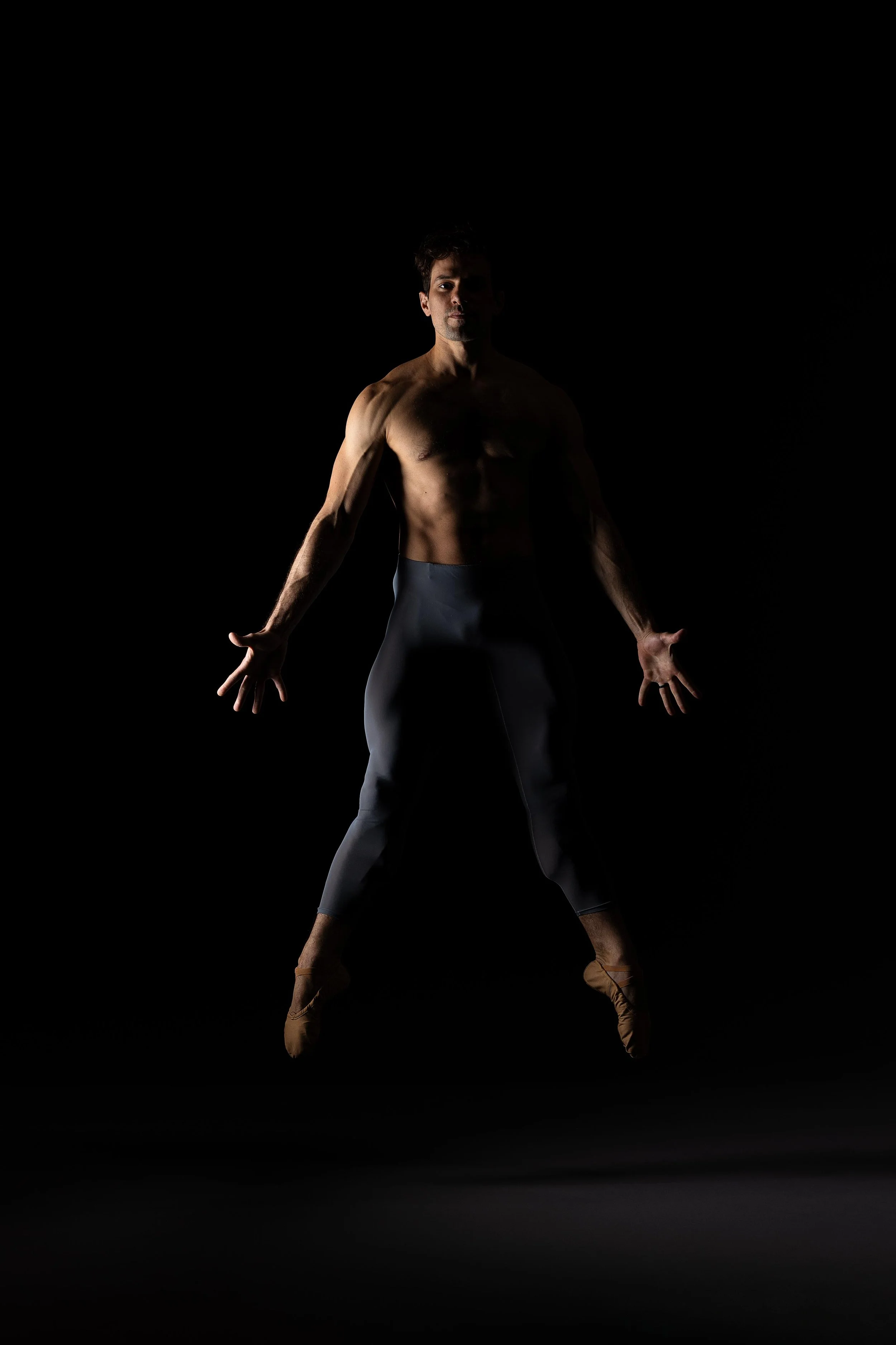 A male ballet dancer in mid-air pose, shirtless, wearing dark pants and pointe shoes, against a black background.