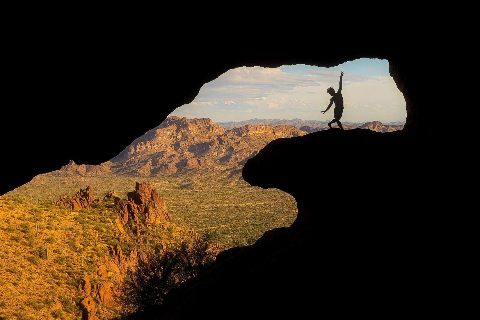 Person standing on rock inside a cave, silhouetted against a desert landscape with mountains and a partly cloudy sky.