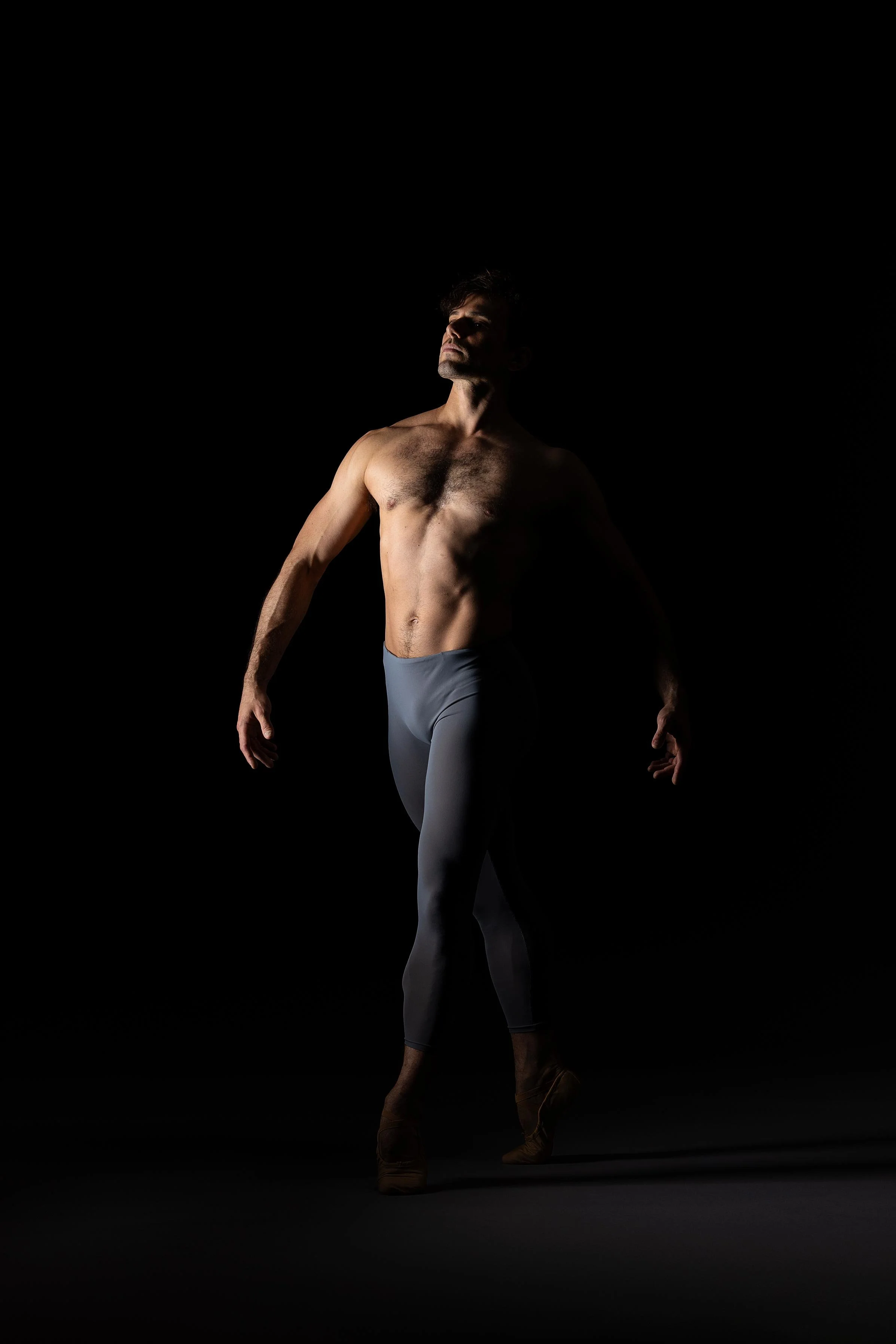 A shirtless male ballet dancer in gray tights and ballet shoes performs against a black background, illuminated by dramatic lighting.