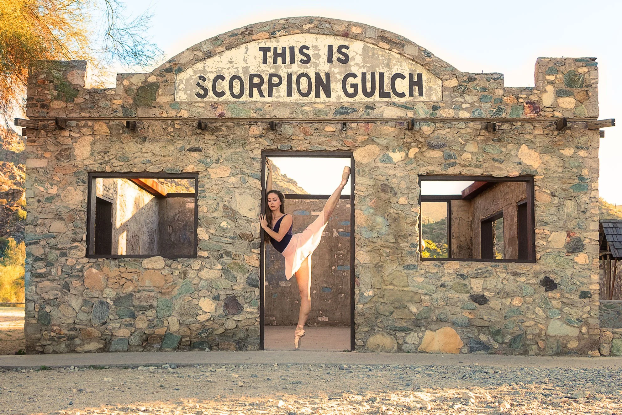 A ballerina practicing in front of a stone building with a sign that reads "This is Scorpion Gulch" during sunset.
