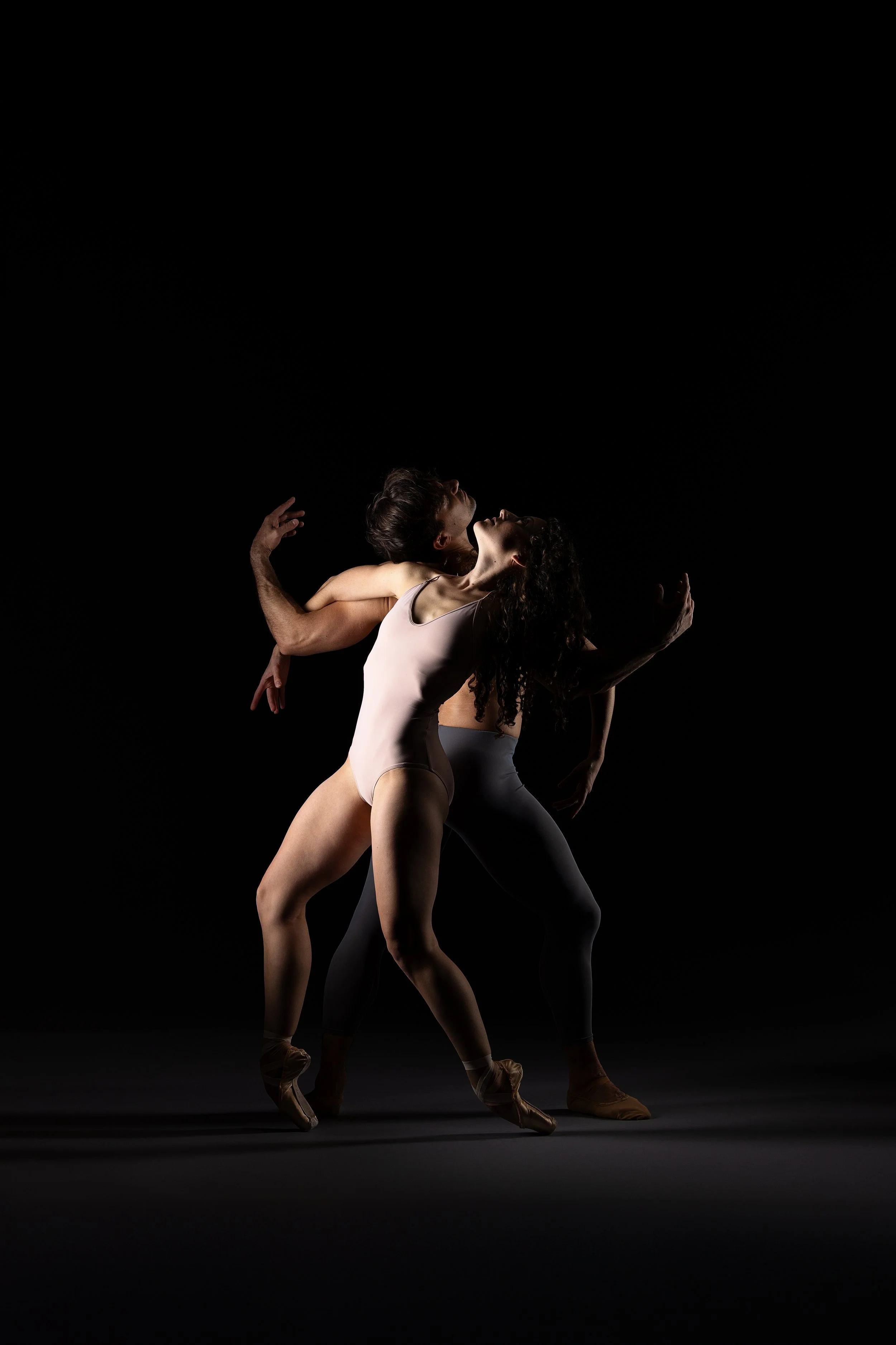 Two ballet dancers performing on a dark stage, one in a light costume and the other in darker attire, captured mid-movement with expressive poses.