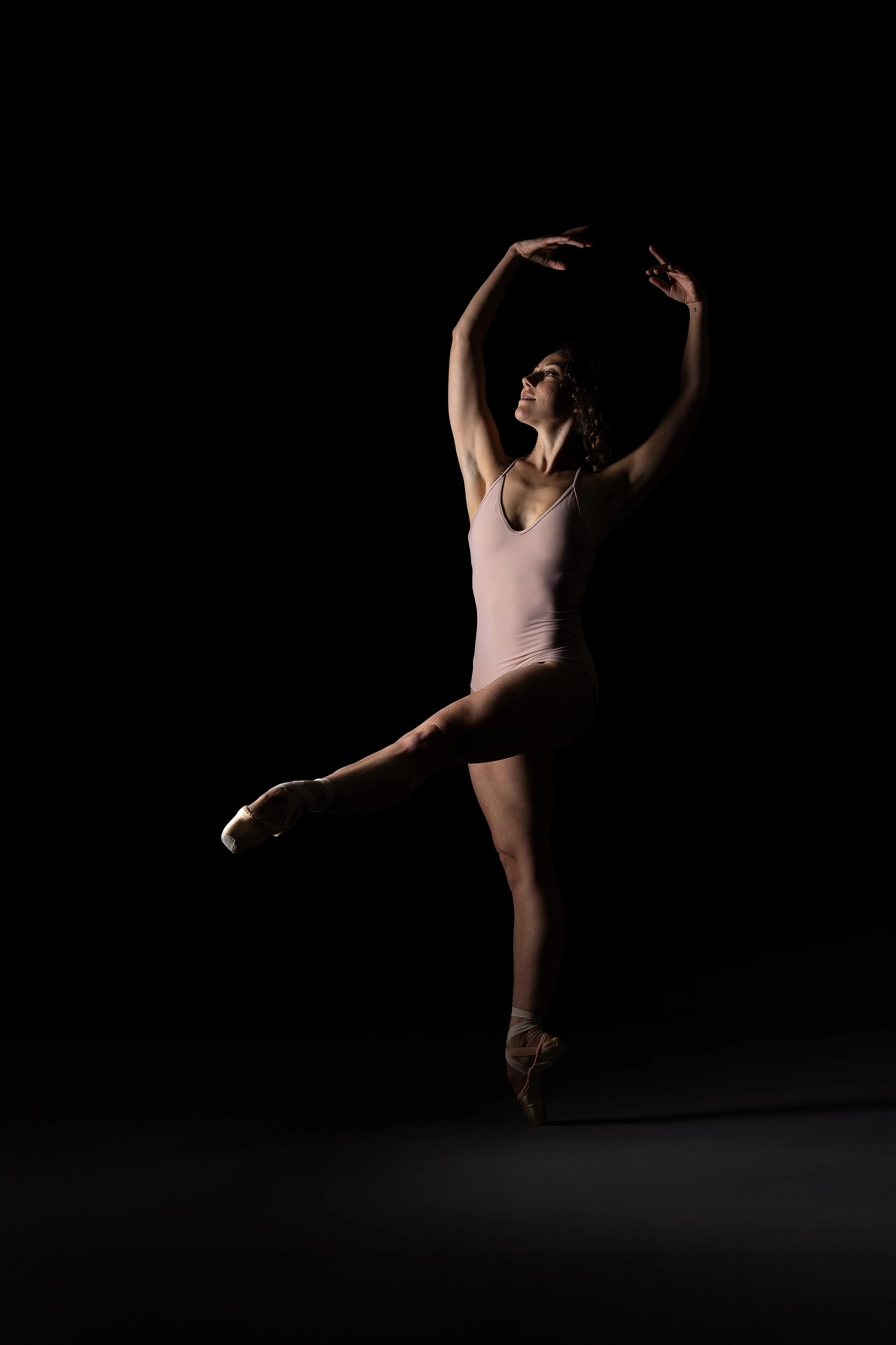 Ballet dancer in a pink leotard performing a pose on pointe against a black background.