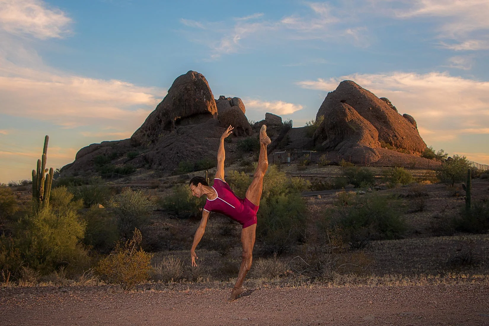 A man performing a dance pose or gymnast move on a desert landscape with large rock formations and cacti during sunset.