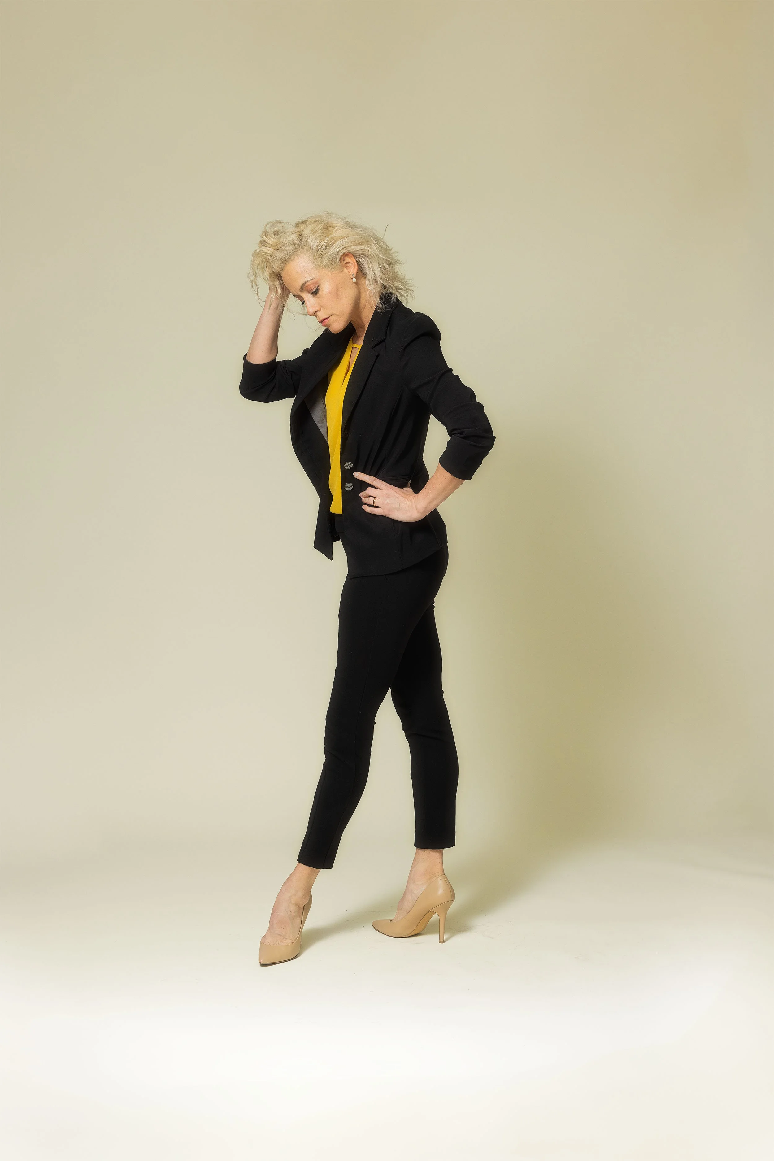 A woman with curly blonde hair wearing a black blazer, yellow top, black pants, and beige high heels, standing against a plain beige background with one hand on her head and the other on her waist.