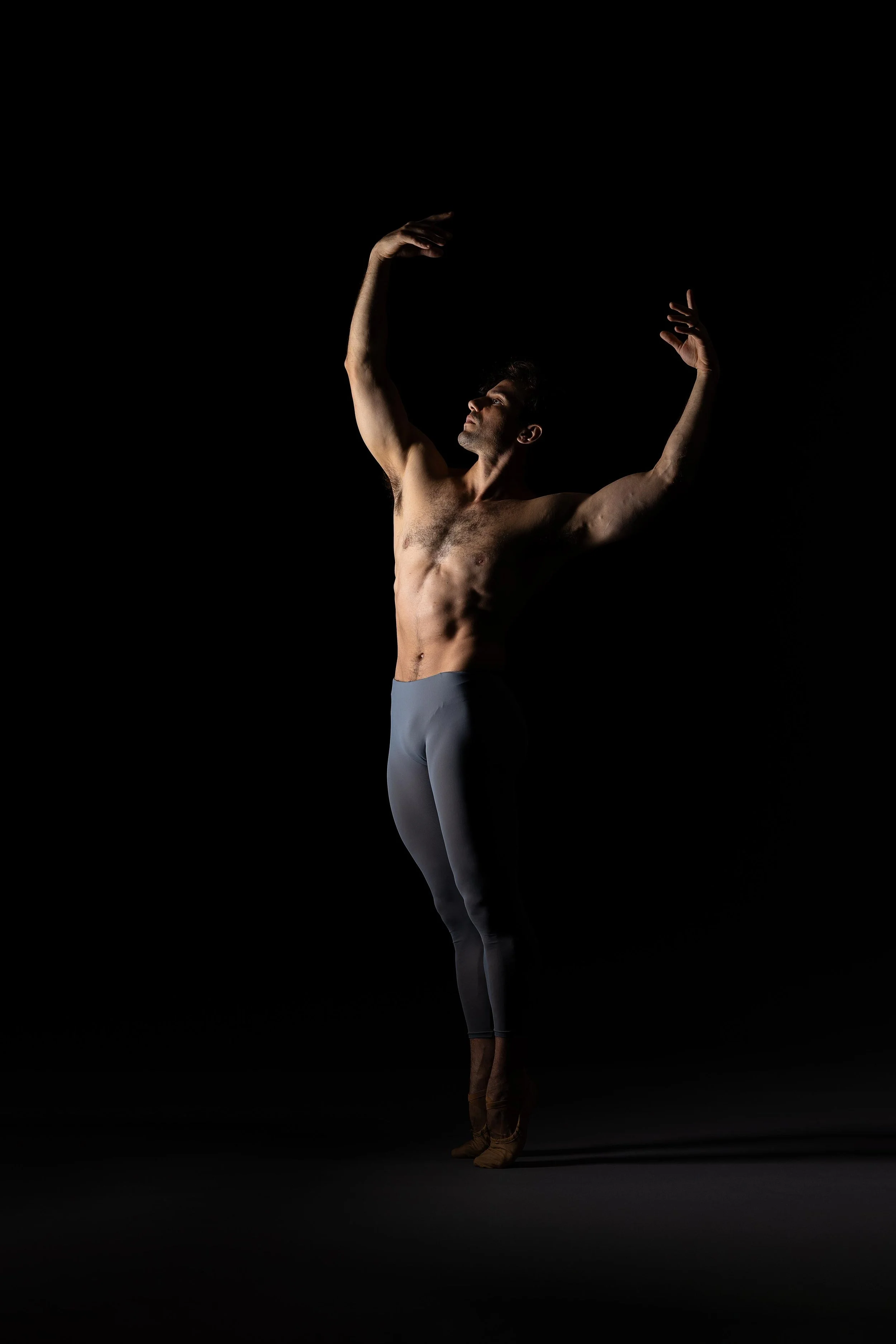A shirtless male ballet dancer in gray tights and brown ballet shoes performing a pose with arms raised against a black background.