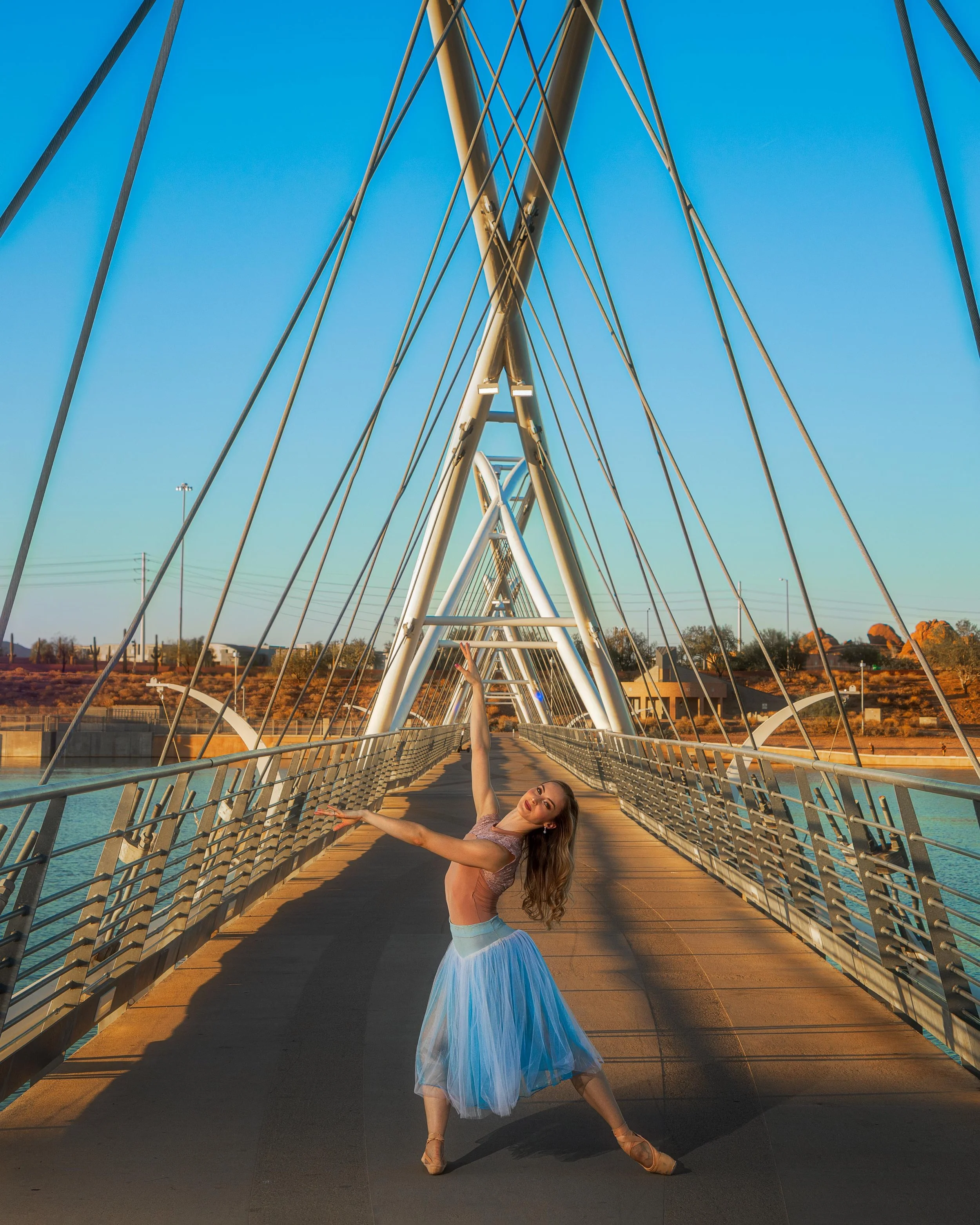 A woman in a pink top and blue tulle skirt dancing on a modern suspension bridge with metal cables and towers, under a clear blue sky.