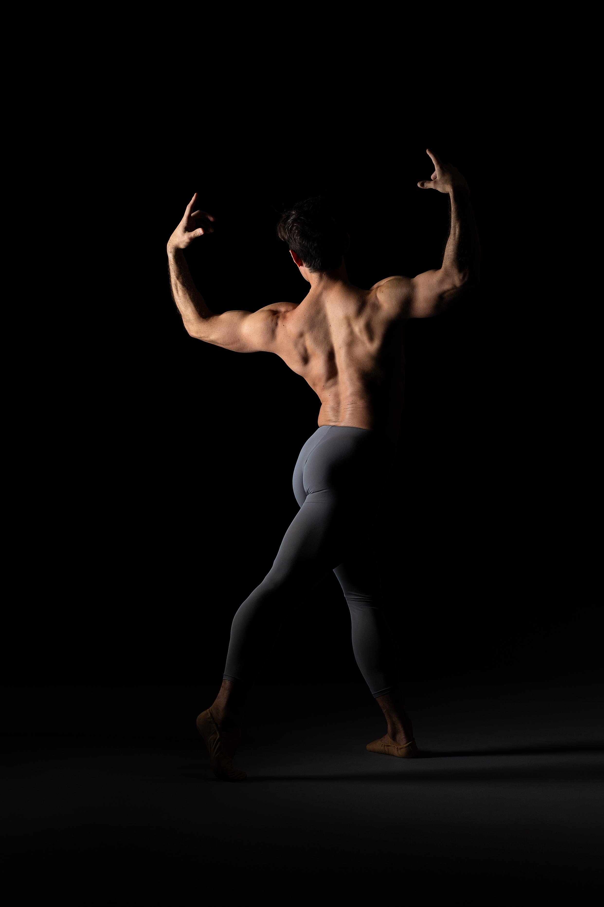 A shirtless man with muscular arms and back, wearing grey leggings, poses with arms raised and flexed against a black background.