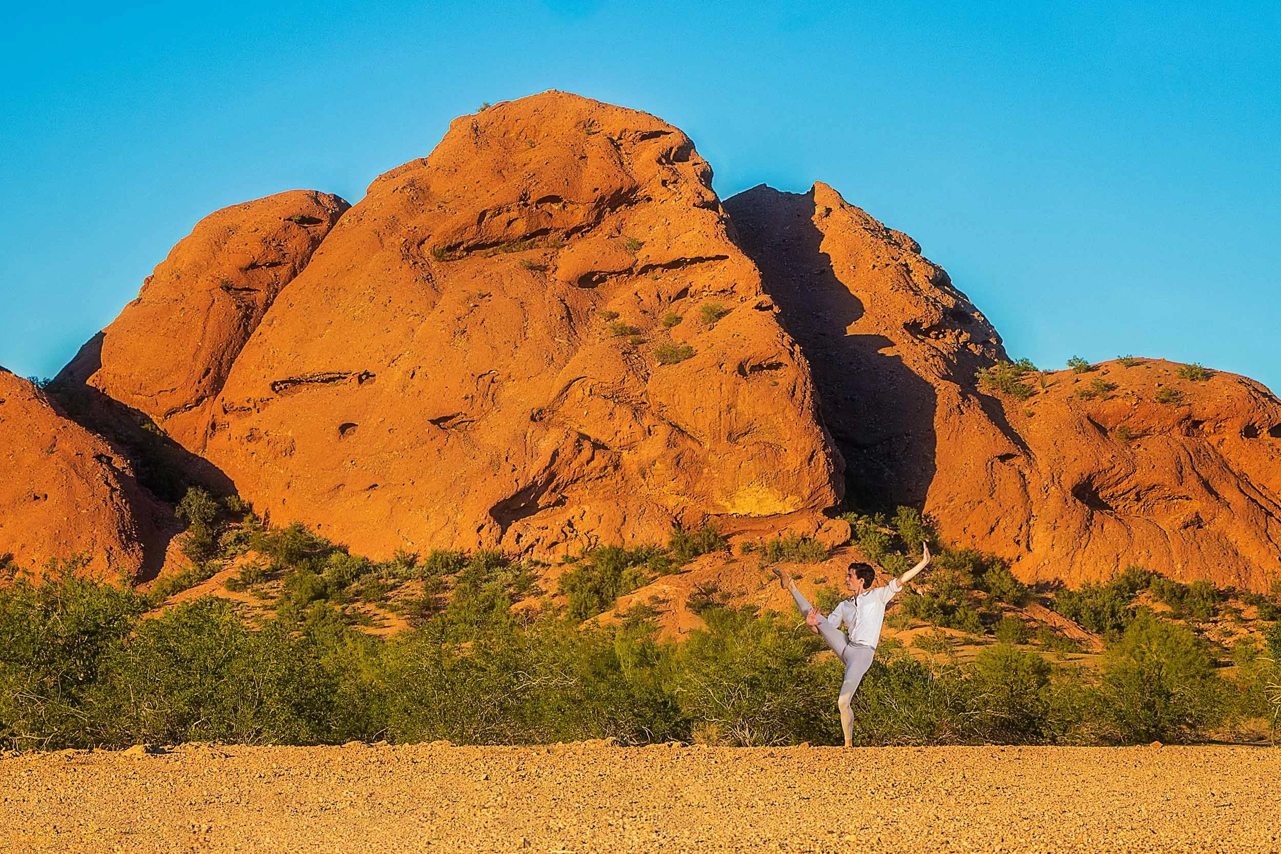 Person practicing yoga in front of a large red rock formation and green bushes on a desert landscape during sunset.