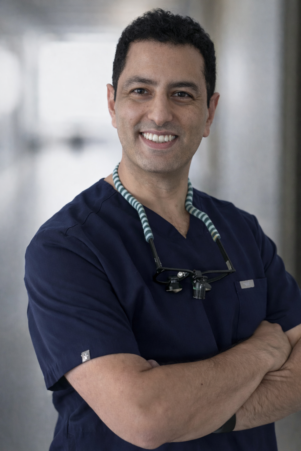A smiling male healthcare professional in navy scrubs with a stethoscope around his neck, standing with arms crossed in a hospital corridor.