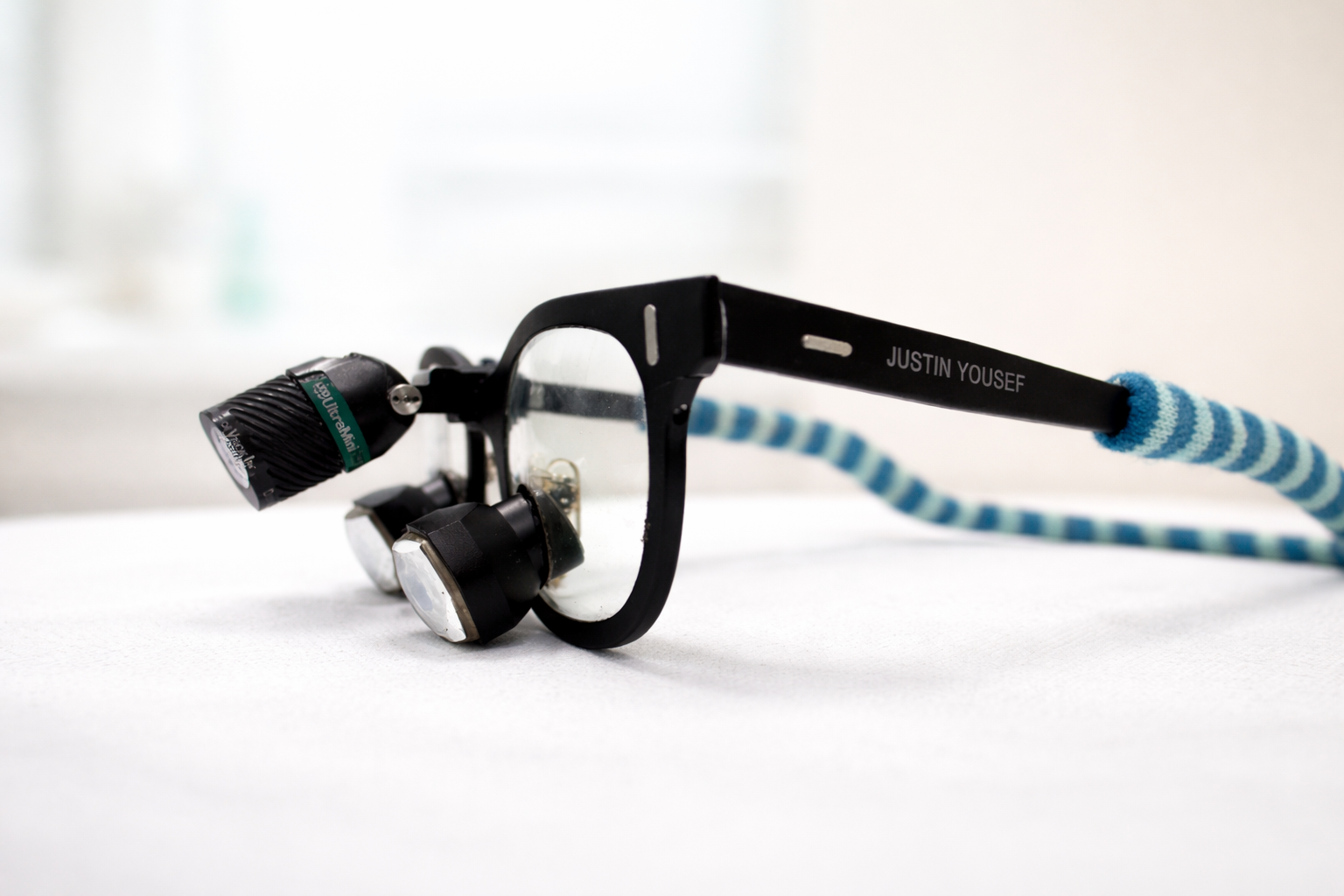 A pair of black eyeglasses with a blue striped head strap attached to the temples, resting on a white surface, with a small black camera attached to one temple.