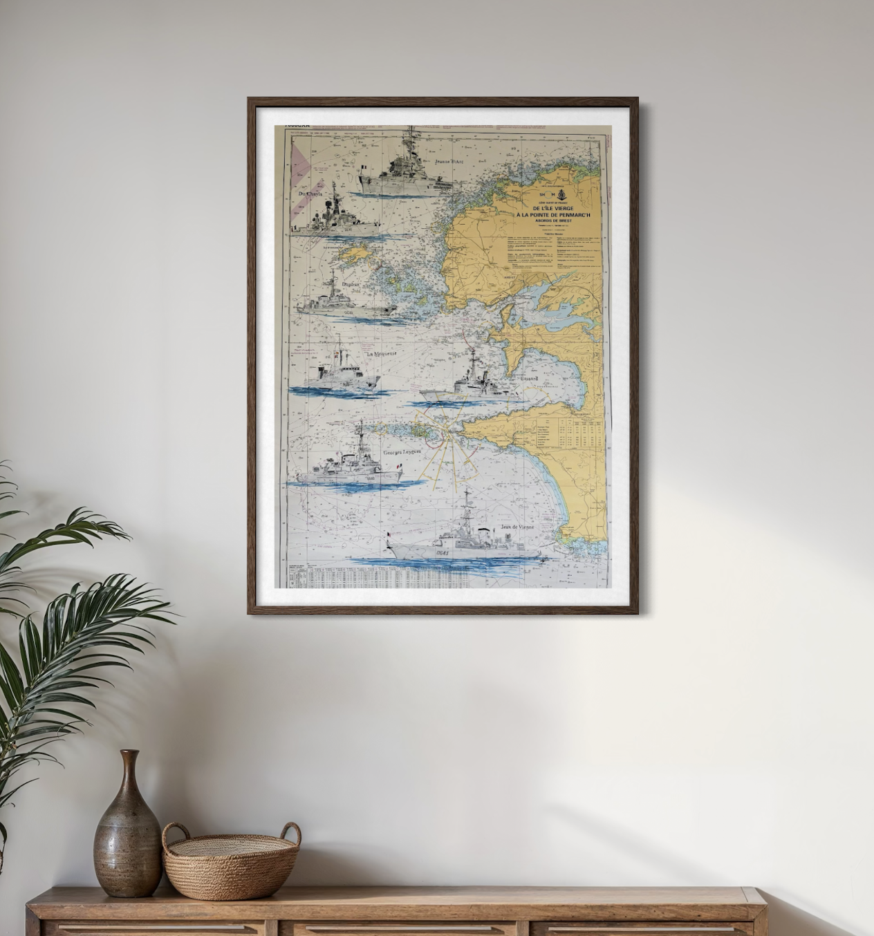 Carte maritime encadrée suspendue sur un mur blanc, avec une table en bois en dessous, décorée de deux vases et d'un panier.