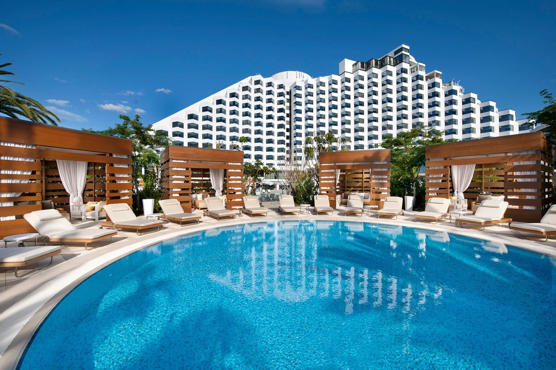 Crown Metropol swimming pool redevelopment in PERTH WA. Outdoor hotel pool area with lounge chairs and cabanas, with a large modern white hotel building in the background under a clear blue sky.
