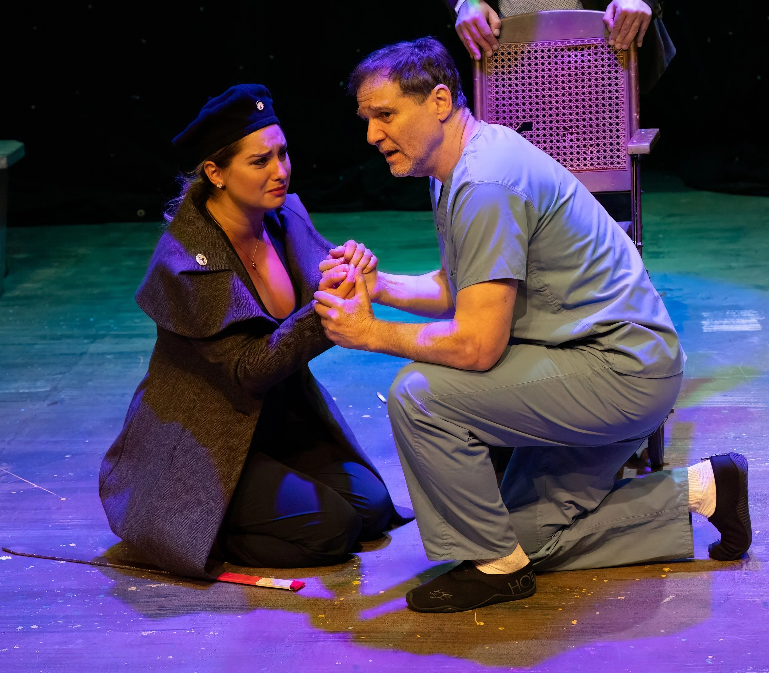 Two actors on stage. One woman kneels, hands clasped with a man who is also kneeling, holding her hands. The woman appears distressed, the man appears earnest. There is a chair behind them and a portion of a third person visible standing.