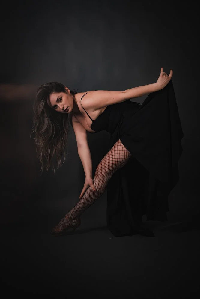 Woman in a black dress with fishnet stockings posing dramatic against a dark background.