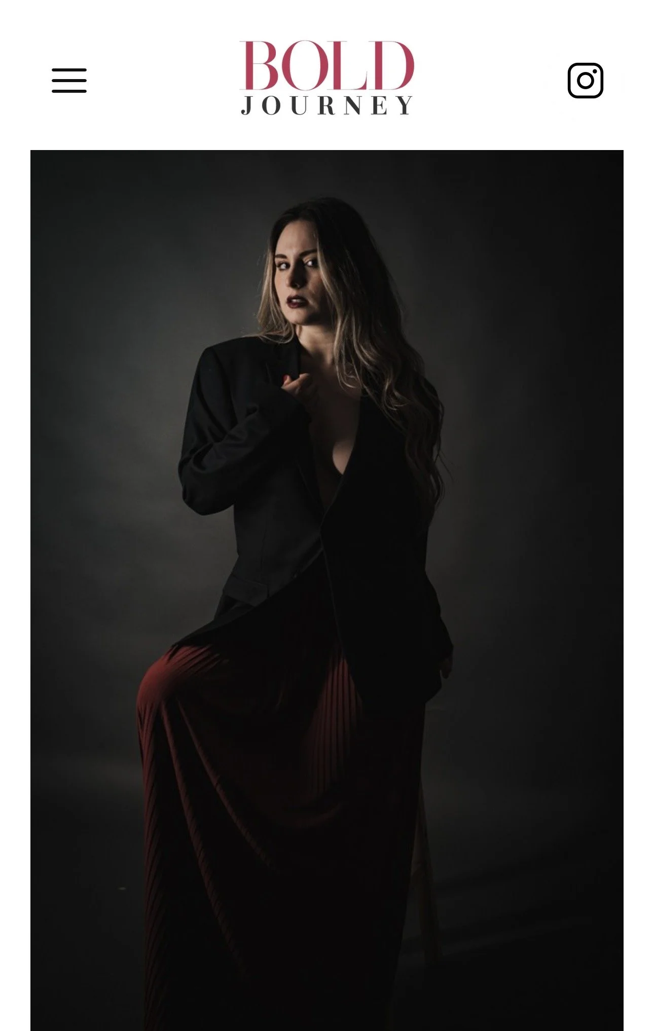 A woman with long wavy hair, dark lipstick, in a black blazer and flowing maroon skirt, standing against a dark gray background.
