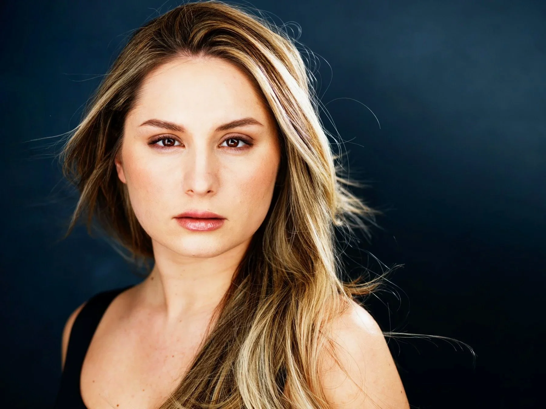 Close-up portrait of a woman with long, wavy blonde hair, wearing a black top, against a dark background.