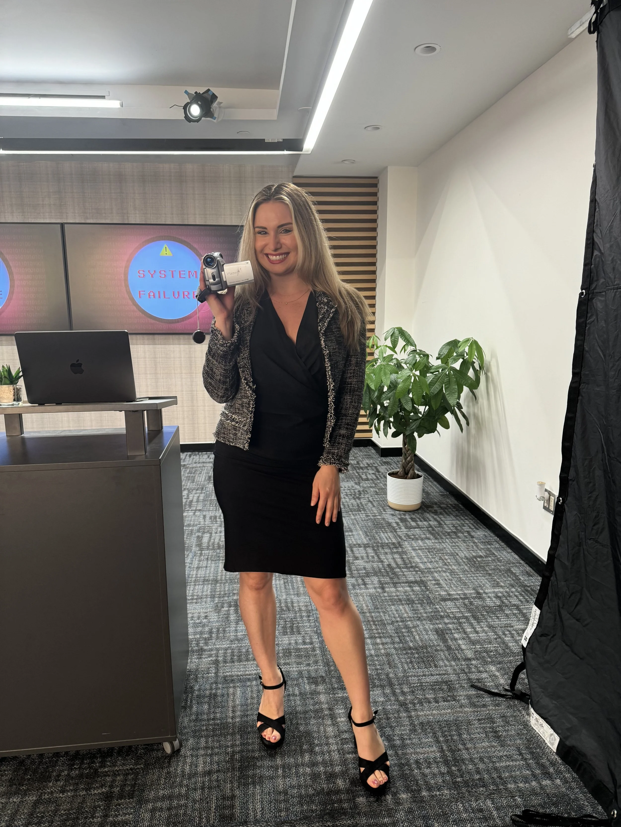 A woman in professional attire holding a video camera with a 'System Failure' warning on the screen behind her in an office environment.