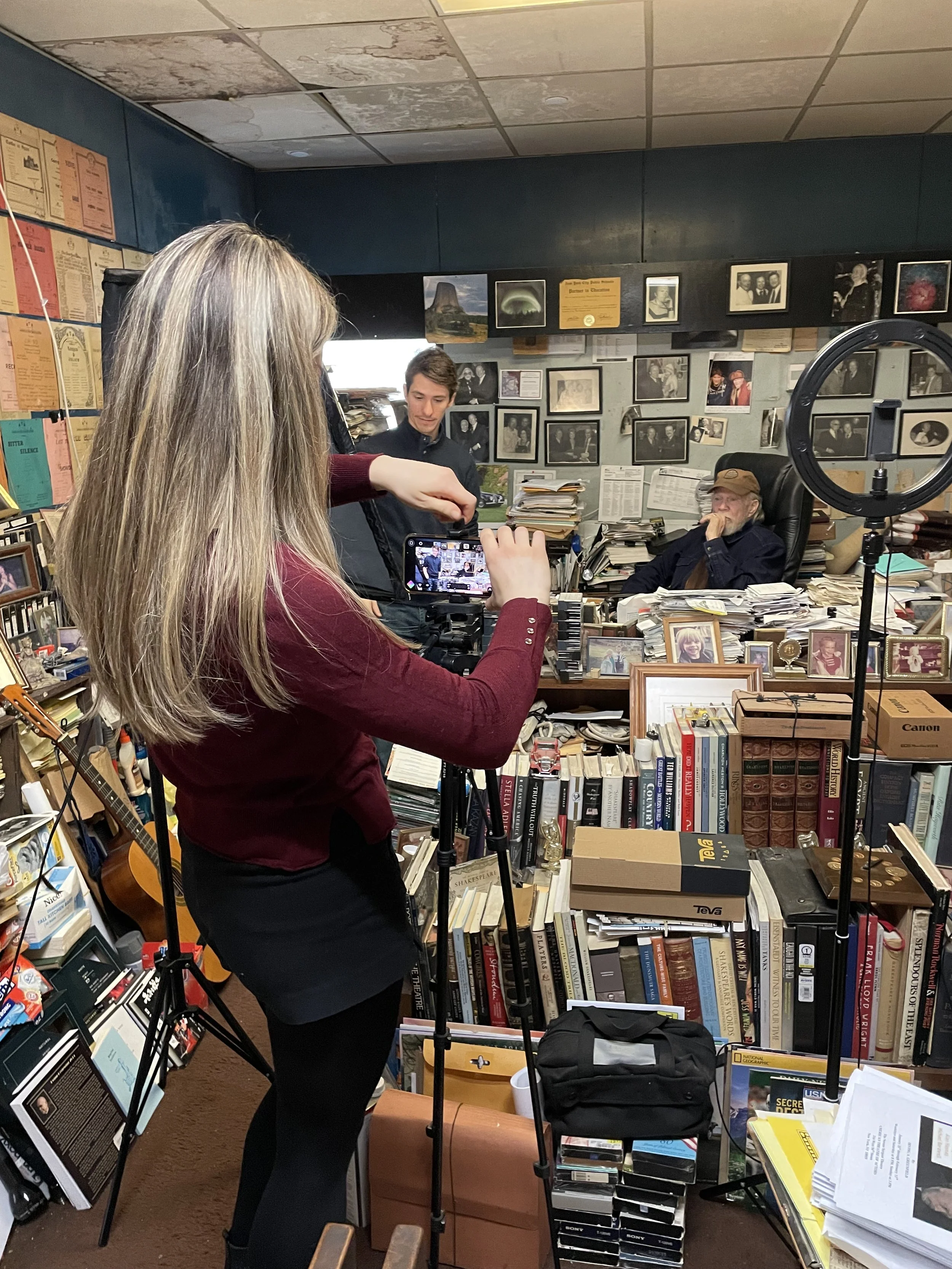 A woman records a man sitting at a cluttered desk in a room filled with books, photographs, and memorabilia.