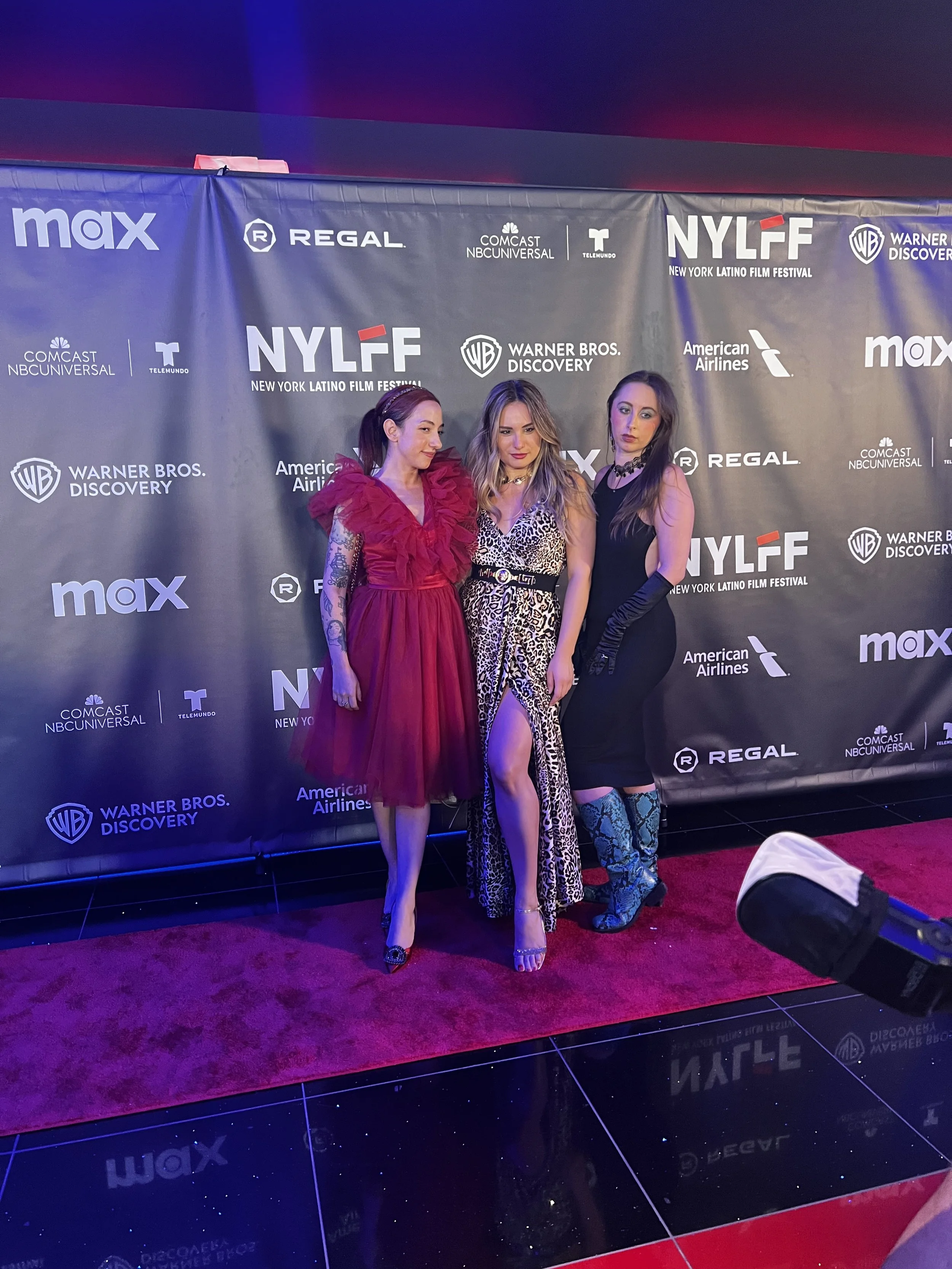 Three women posing on the red carpet at the NYLFF film festival, with a backdrop featuring event logos and sponsors.