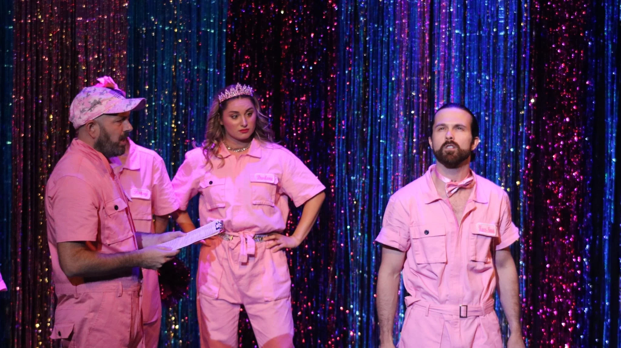 Four performers in pink outfits stand on a stage with colorful, shiny curtain backdrop. One person is reading from a script, the woman is wearing a crown, and the man on the right has a beard and is looking forward.
