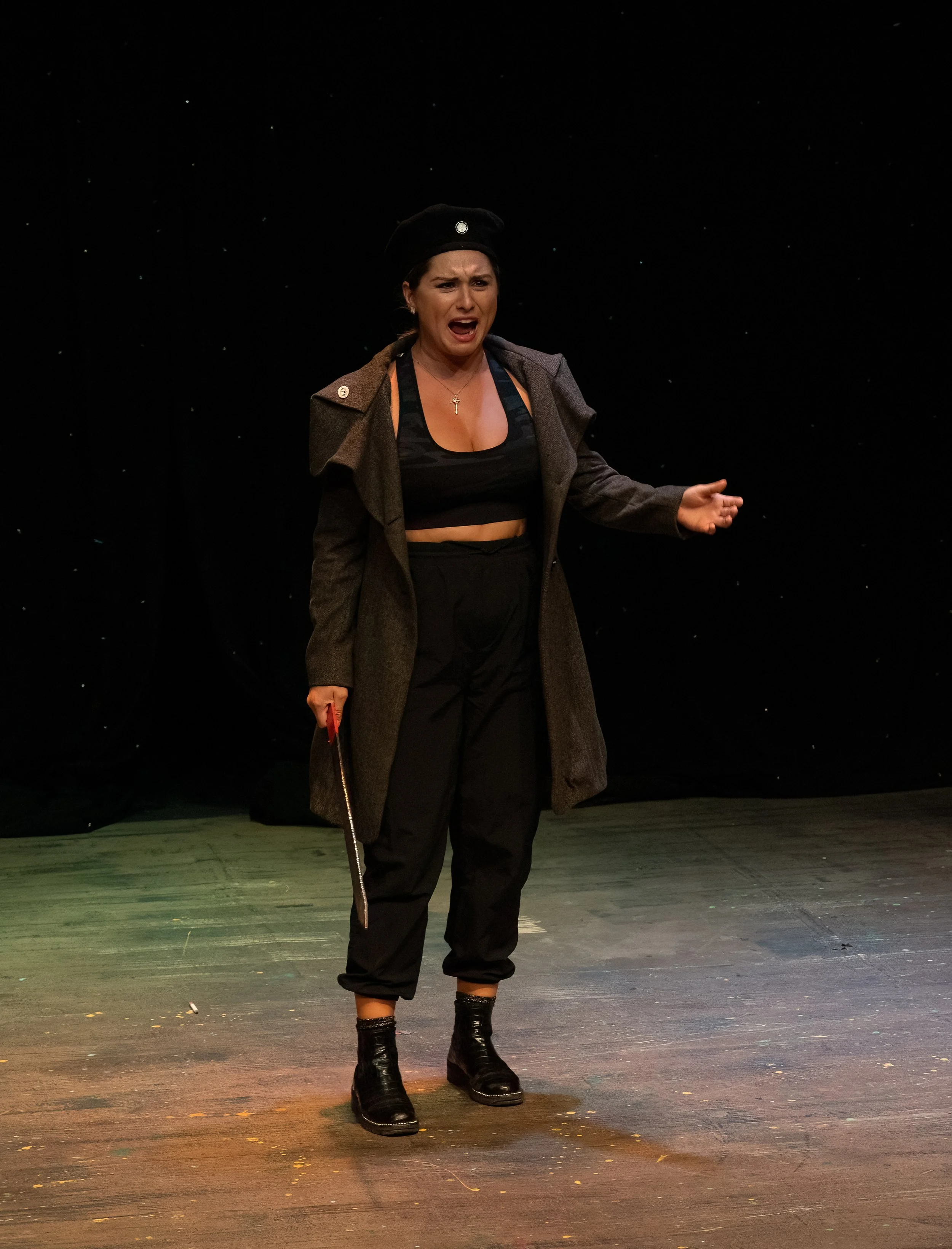 A woman performing on stage, wearing a black hat, a gray coat, black pants, and black boots, with an expressive facial expression.
