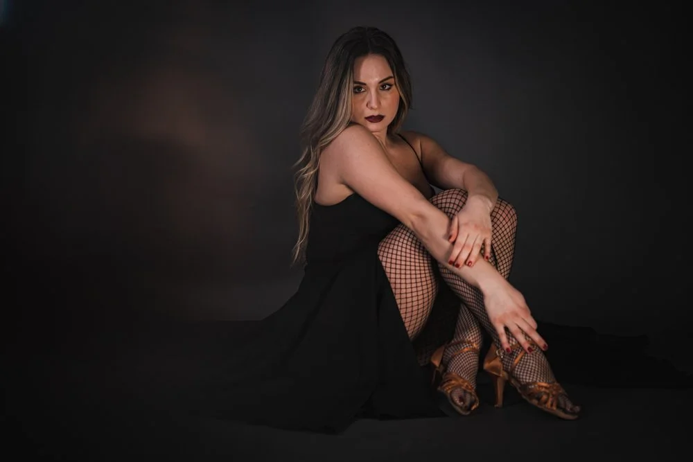 A woman with long, wavy hair, wearing dark lipstick, fishnet stockings, and heels, sitting on the floor with a dark background.