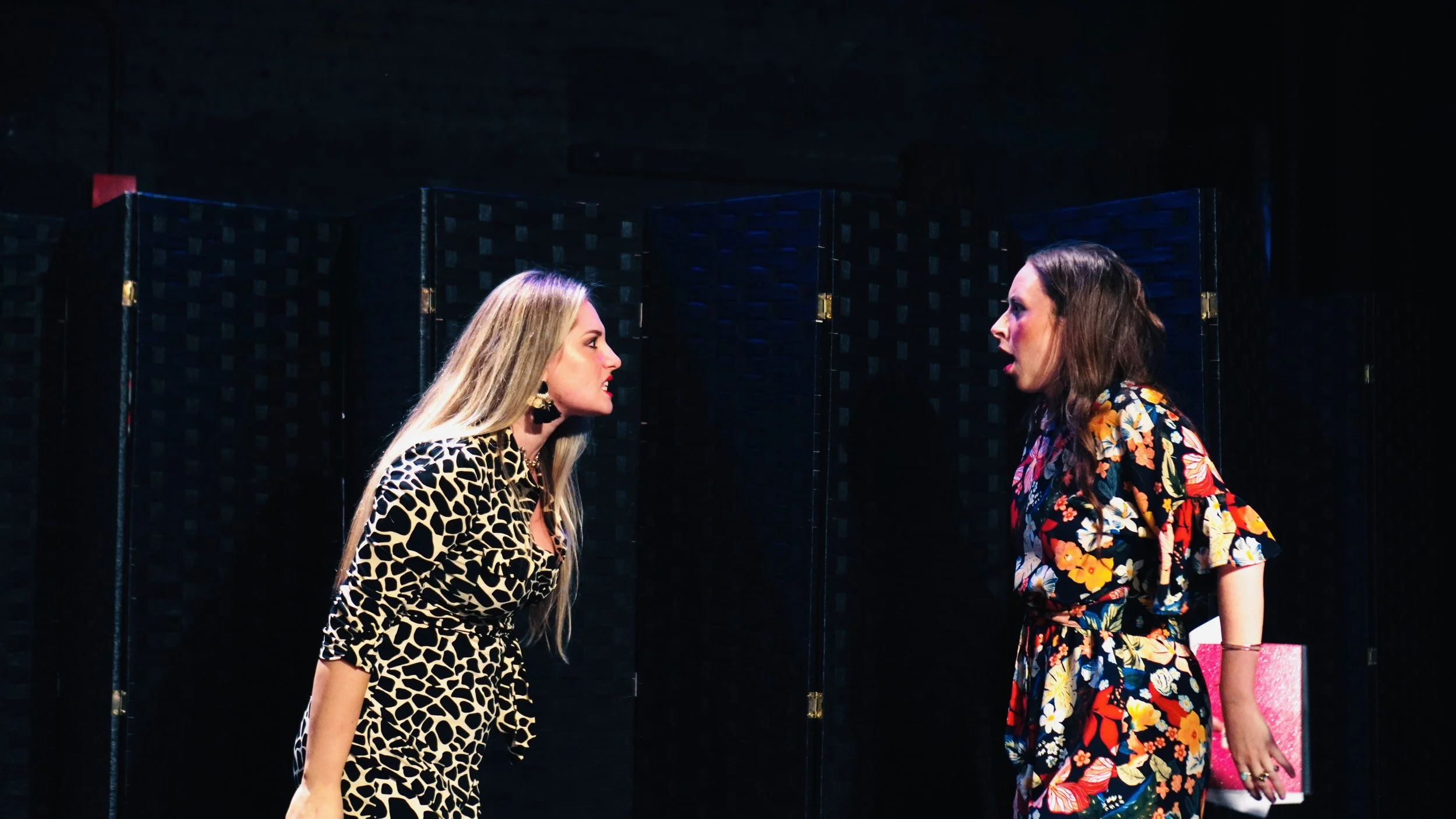 Two women are having an argument on stage, facing each other with serious expressions, with a dark background and a folded screen behind them.