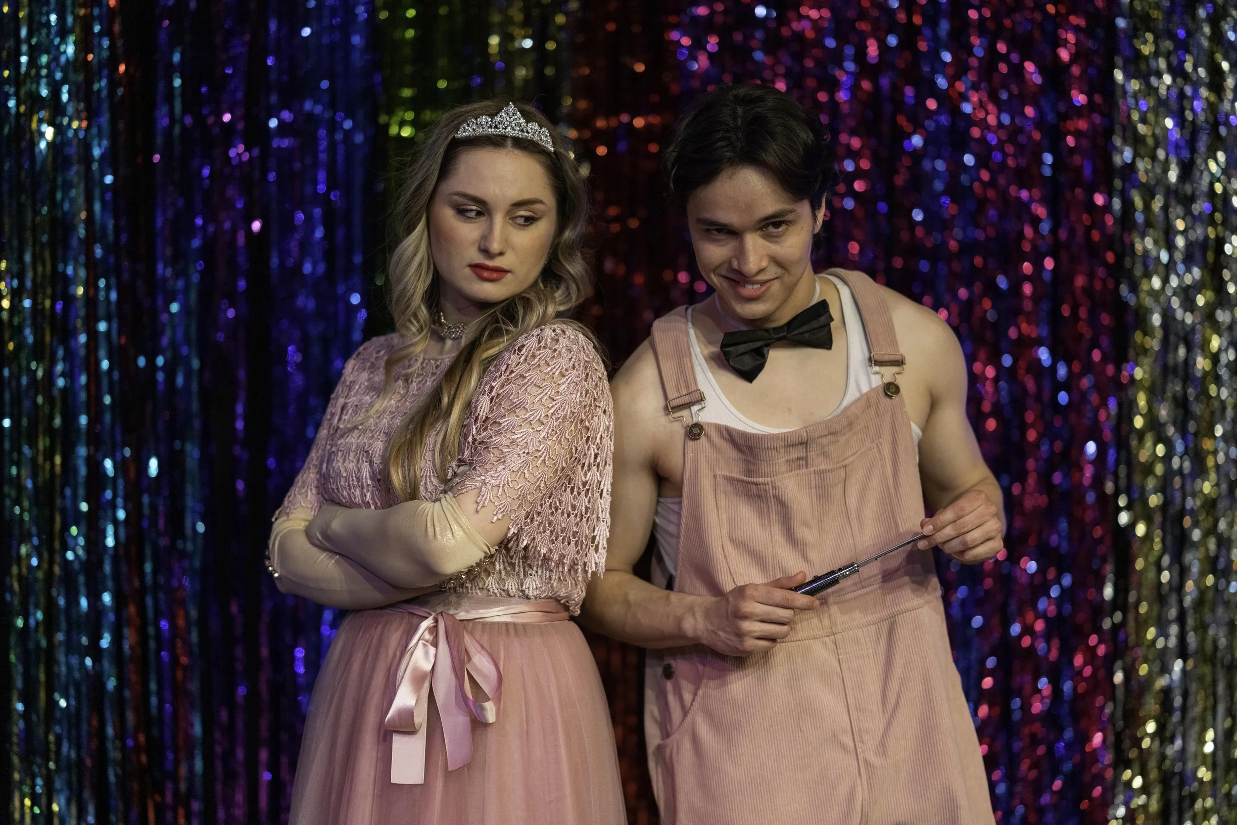 A young woman wearing a tiara and pink dress with lace details, standing next to a young man in pink overalls and a black bow tie, holding a small knife, in front of a colorful glittering curtain background.