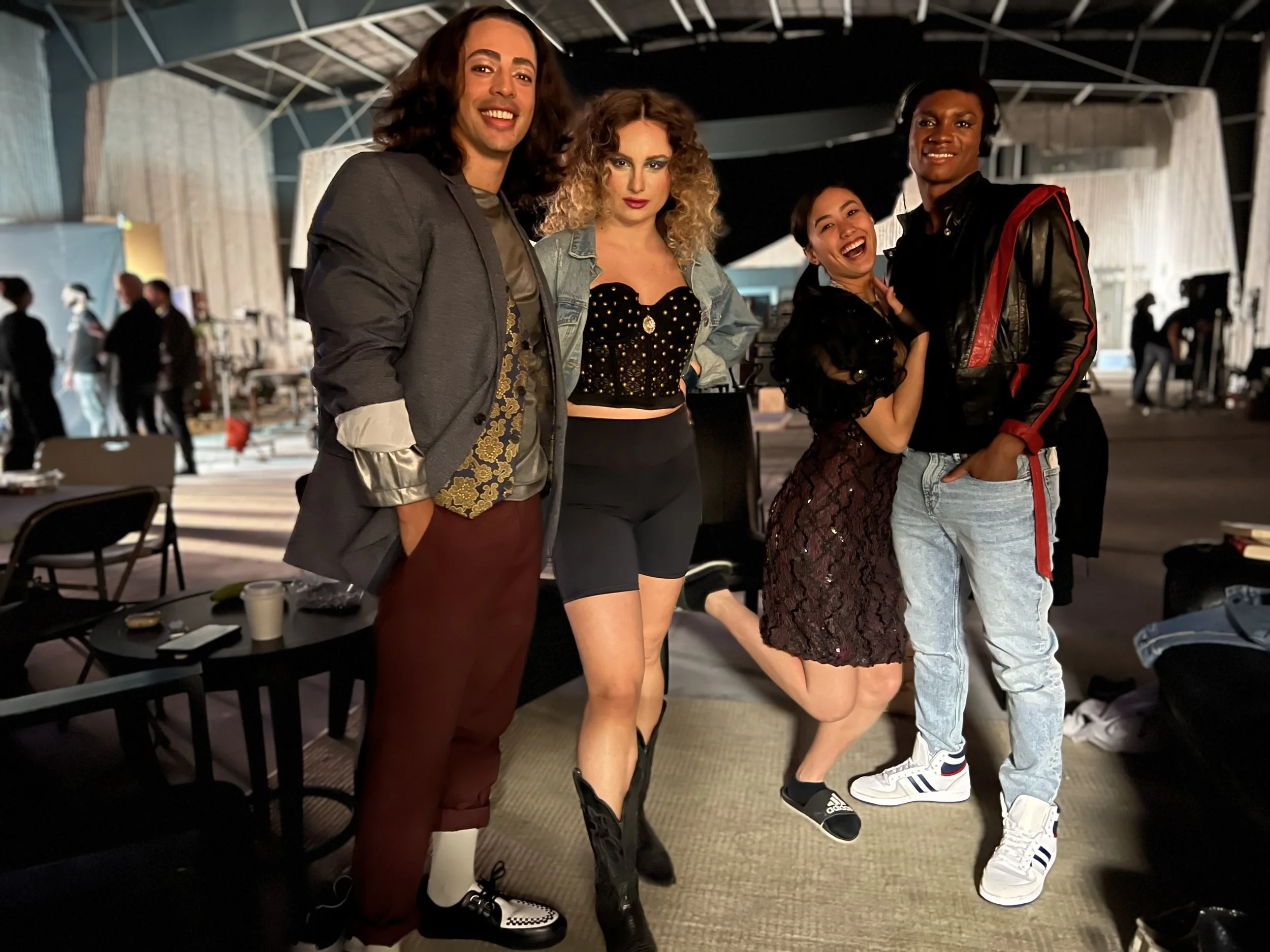 Group of five diverse people standing together backstage, smiling at the camera, with some dressed in casual clothing and others in stylish outfits, and equipment visible in the background.