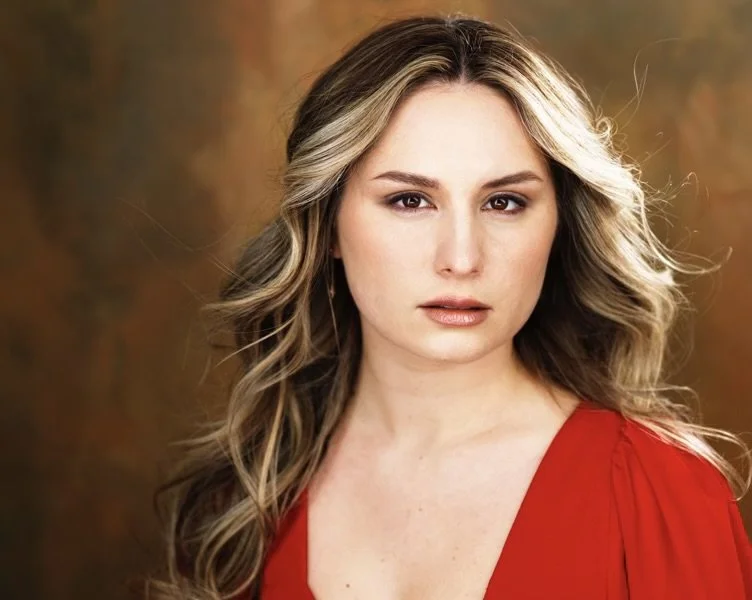 A woman with long, wavy blonde hair and light makeup, wearing a red top, has a serious expression and is looking at the camera against a brown background.
