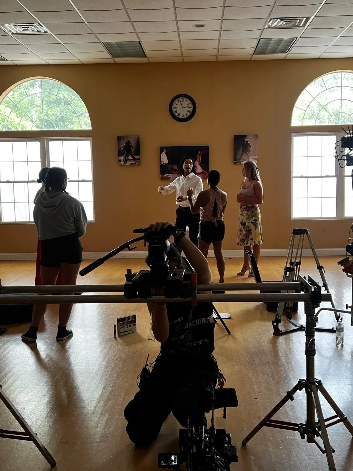 A film crew is filming a scene in a dance studio with four women. One woman is speaking or performing, while others listen. There are large windows, a clock on the wall, and framed photos of dancers.