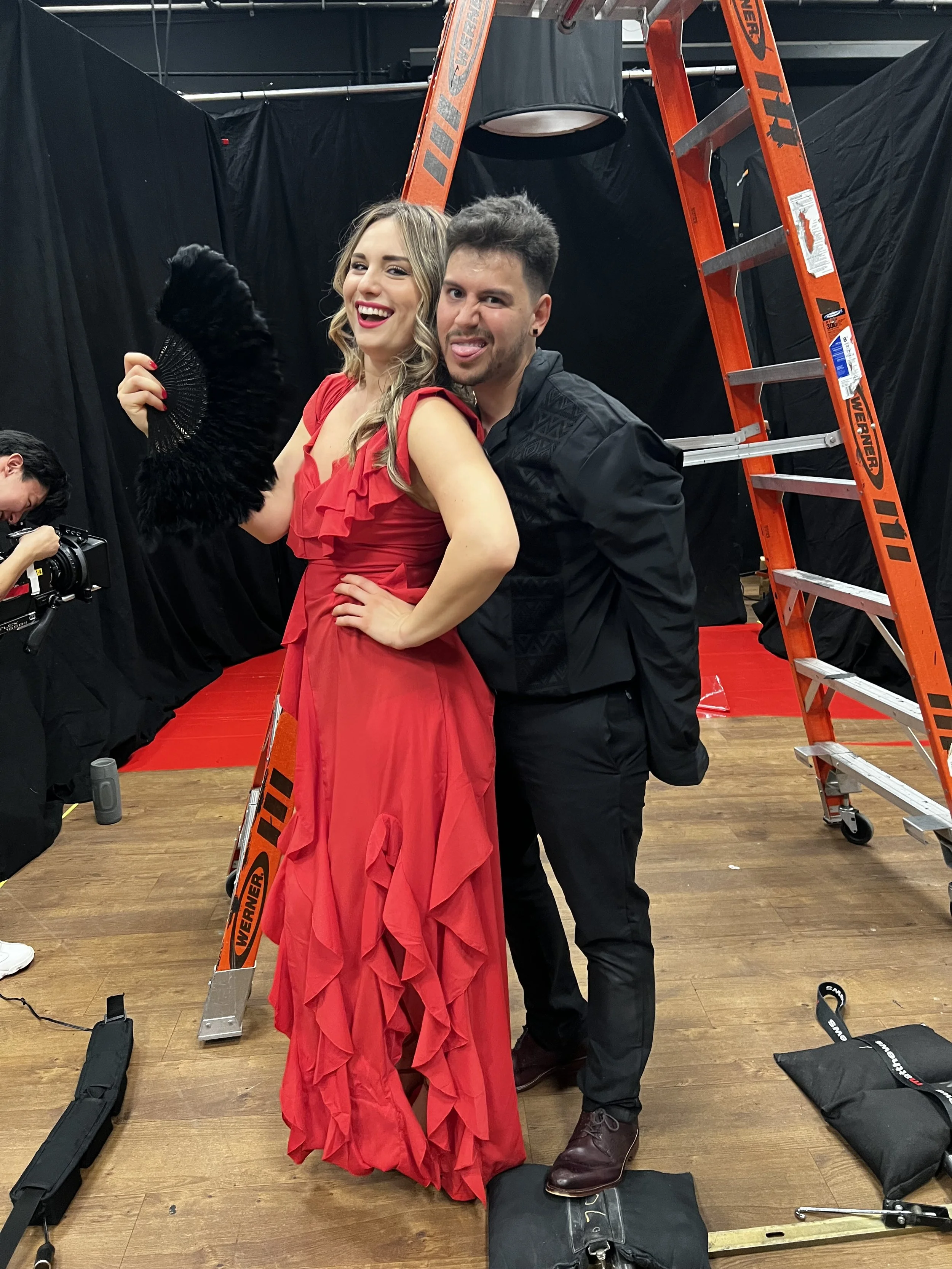 Two people posing in a studio with a ladder and black curtains in the background. The woman is wearing a long red dress and holding a black fan, smiling, and the man is standing close behind her, sticking out his tongue.
