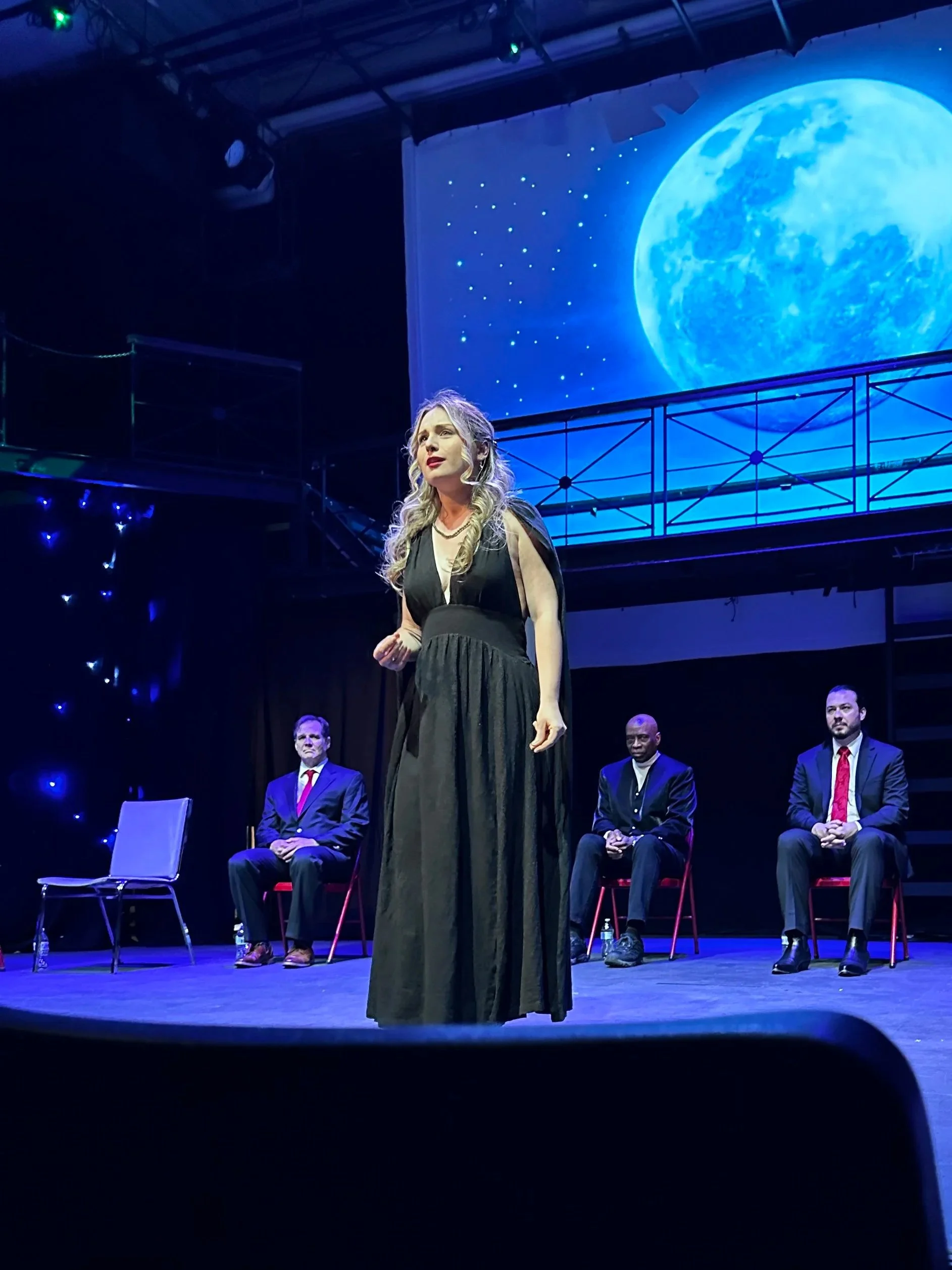 A theatrical performance with four actors on stage, a woman in a black dress standing in front, three men seated behind her, a large blue moon and starry sky on a screen backdrop.