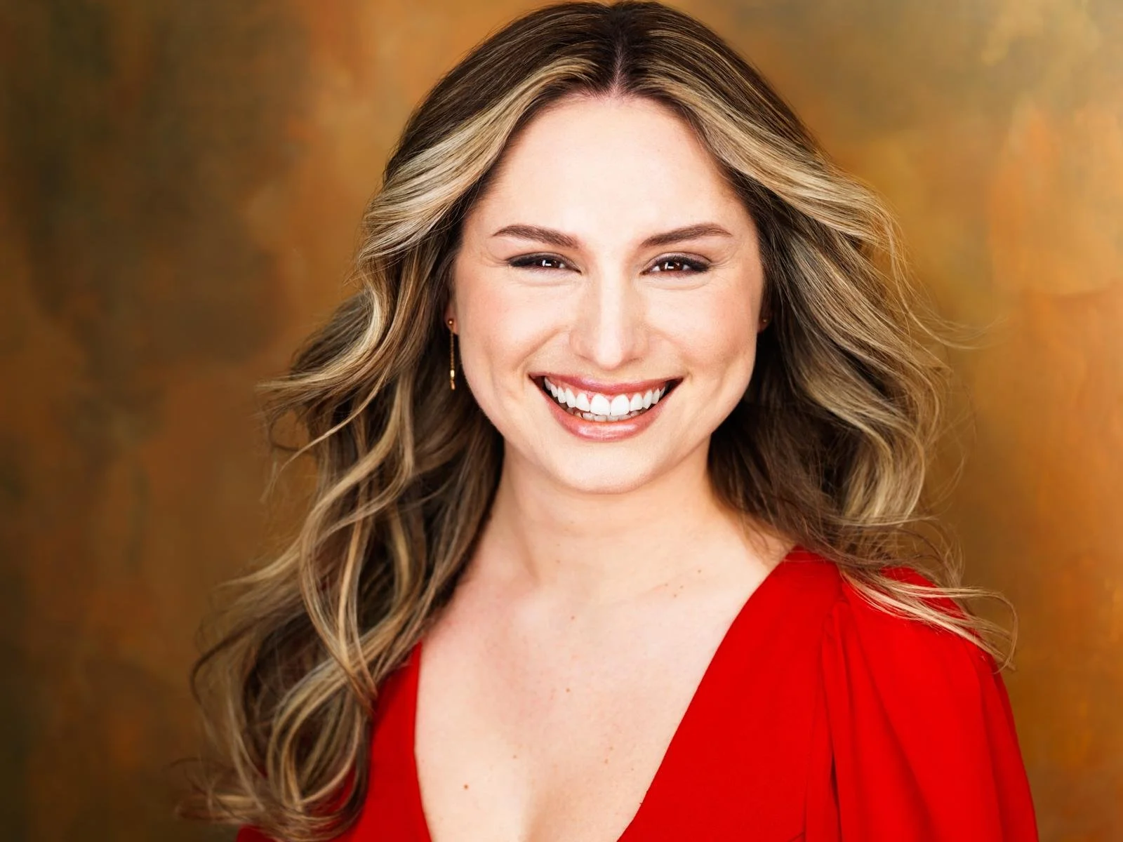 A woman with wavy light brown hair, wearing a red dress, smiling with white teeth, against a warm brown and gold background.