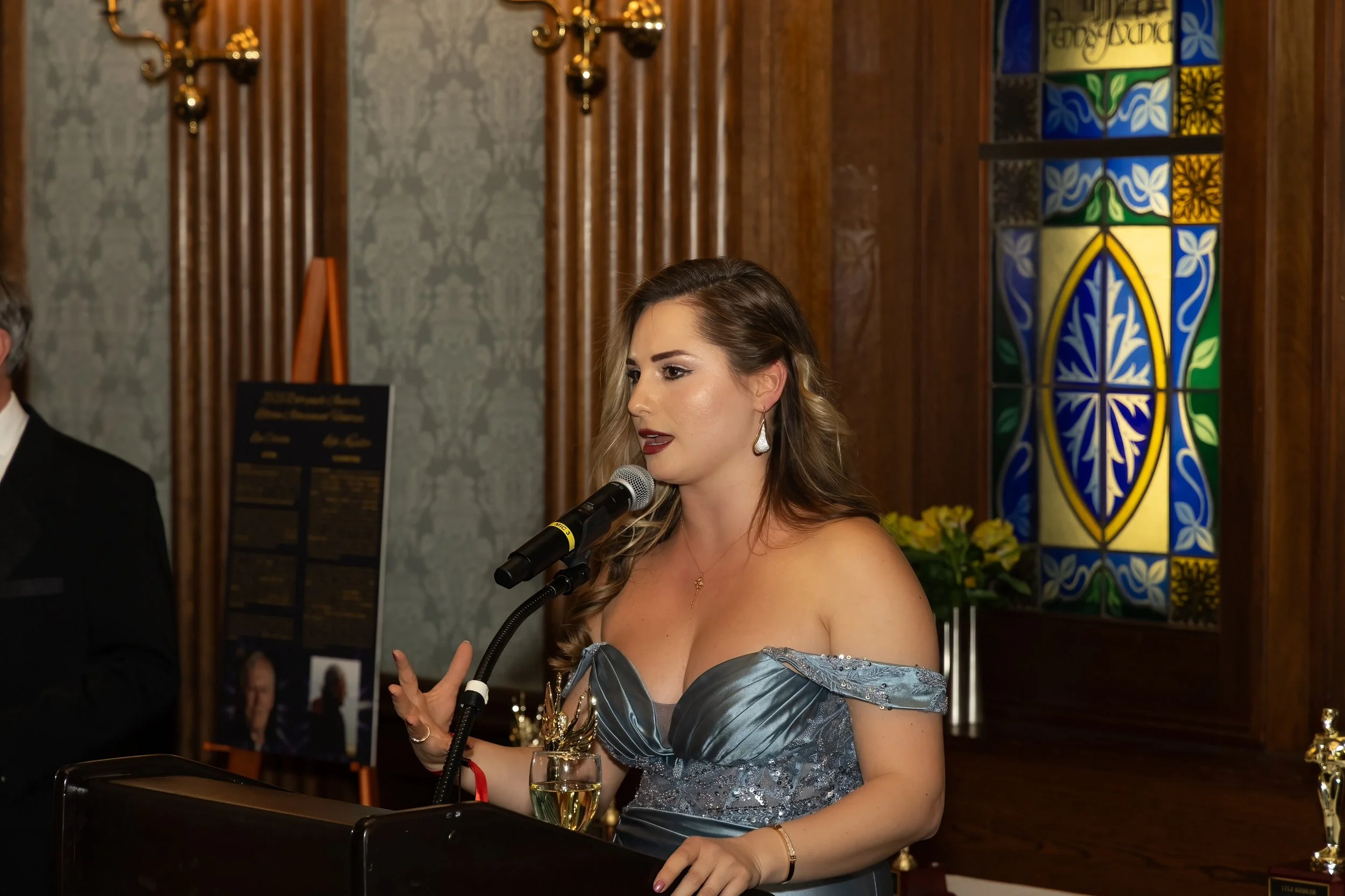 Karolina Larion in an off-the-shoulder dress giving her acceptance speech into a microphone at an award ceremony at the Penn Club in Manhattan.