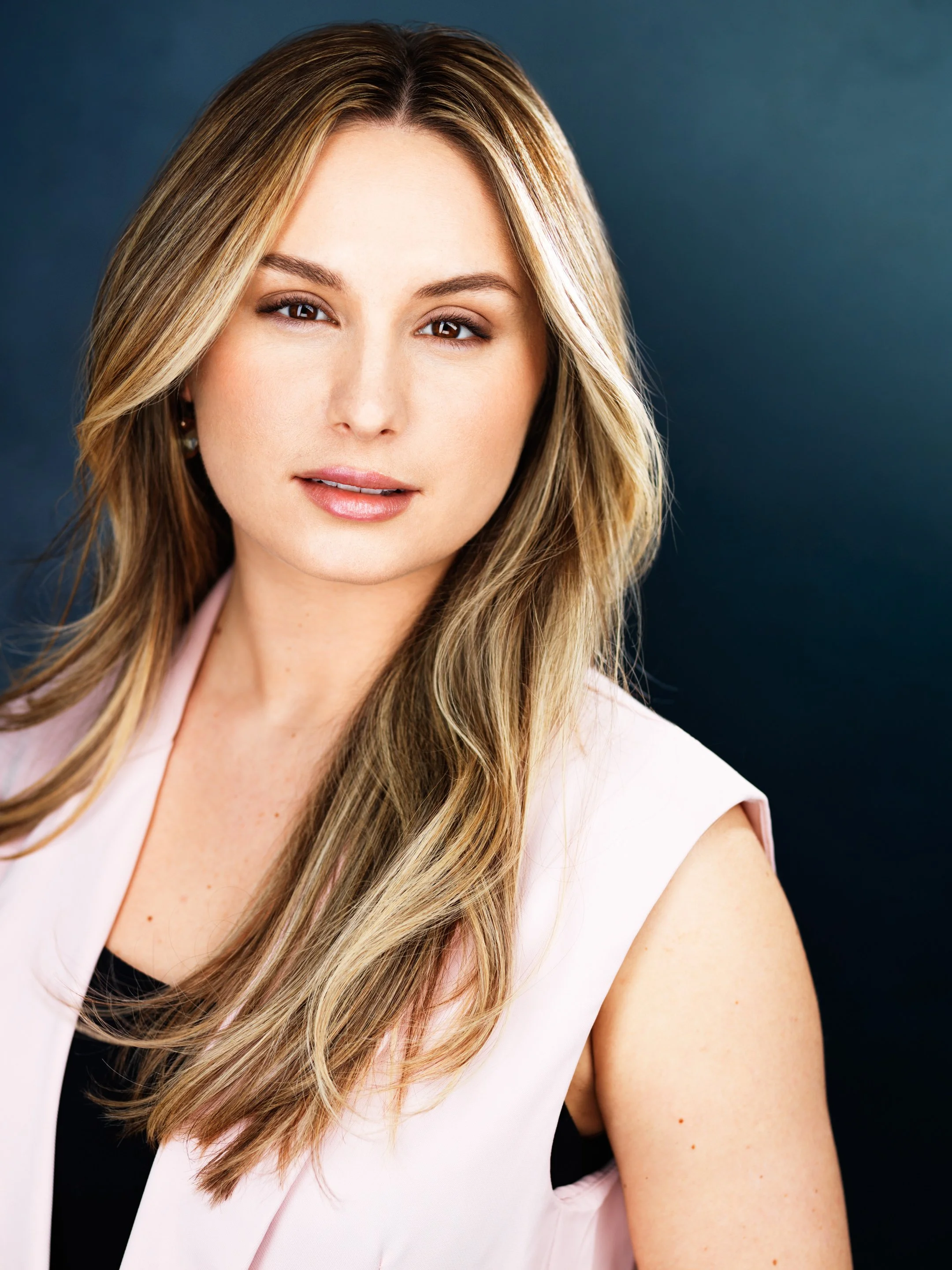 A portrait of a woman with long blonde hair, light makeup, and a neutral expression, wearing a sleeveless light pink top against a dark background.