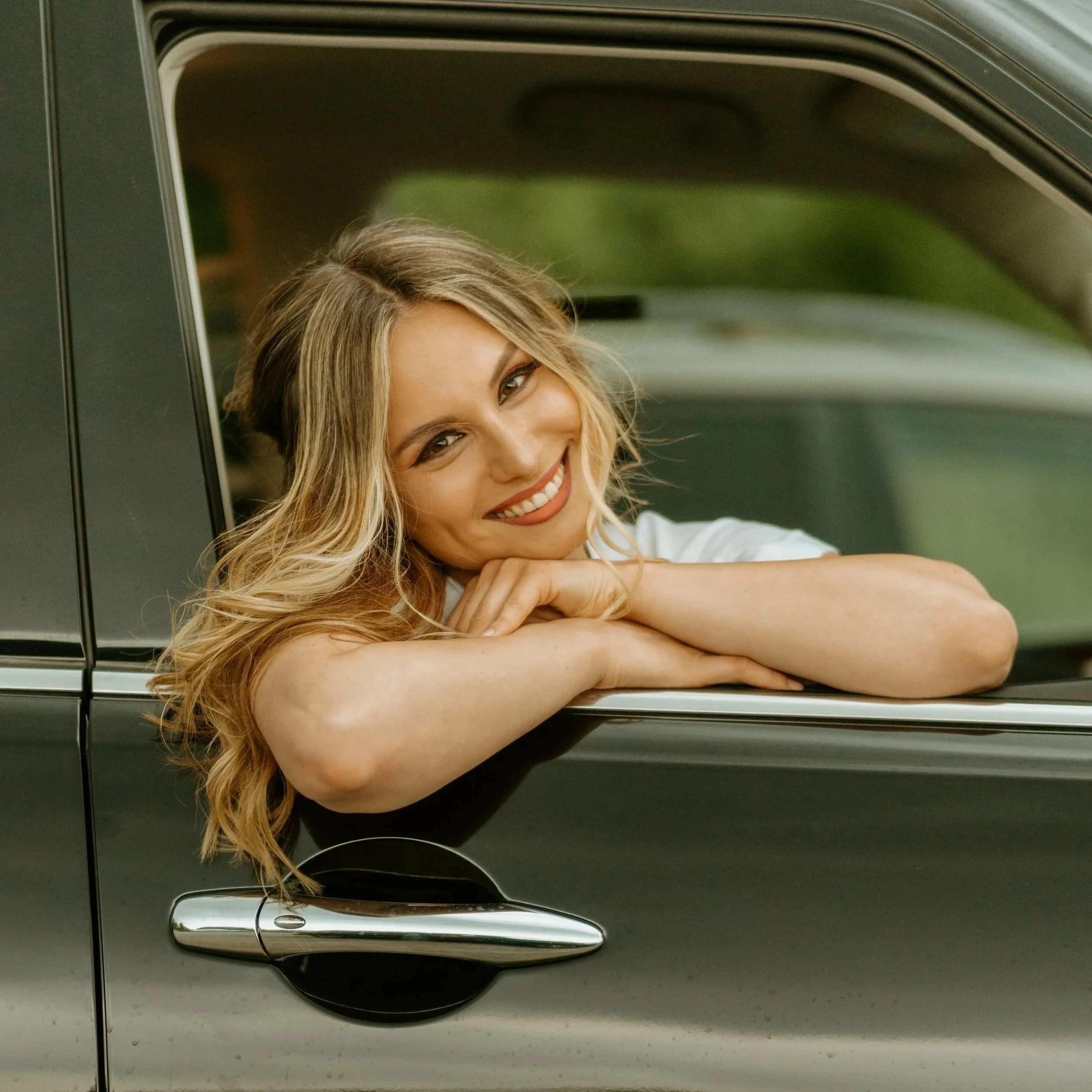 A woman with blonde wavy hair smiling and resting her chin on her crossed arms in a car window.
