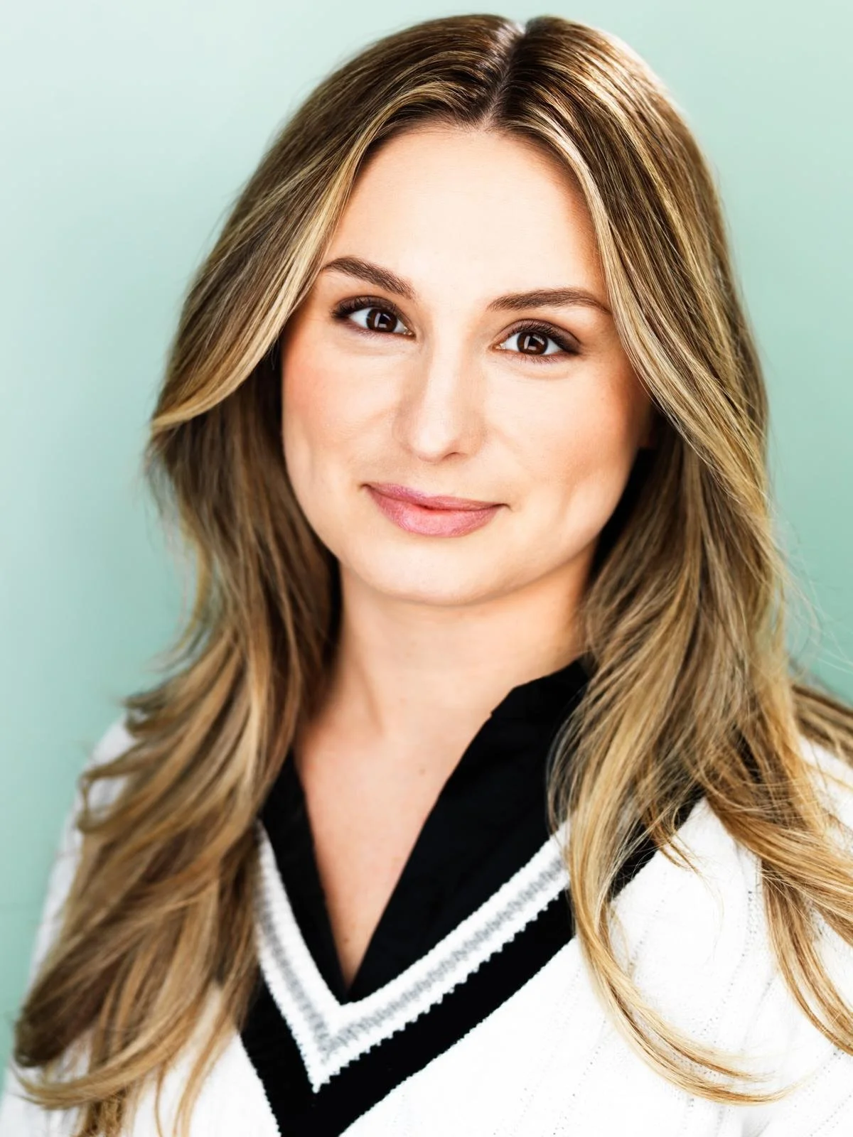 Portrait of a woman with long, light brown hair, smiling, wearing a white sweater with black trim, against a light green background.