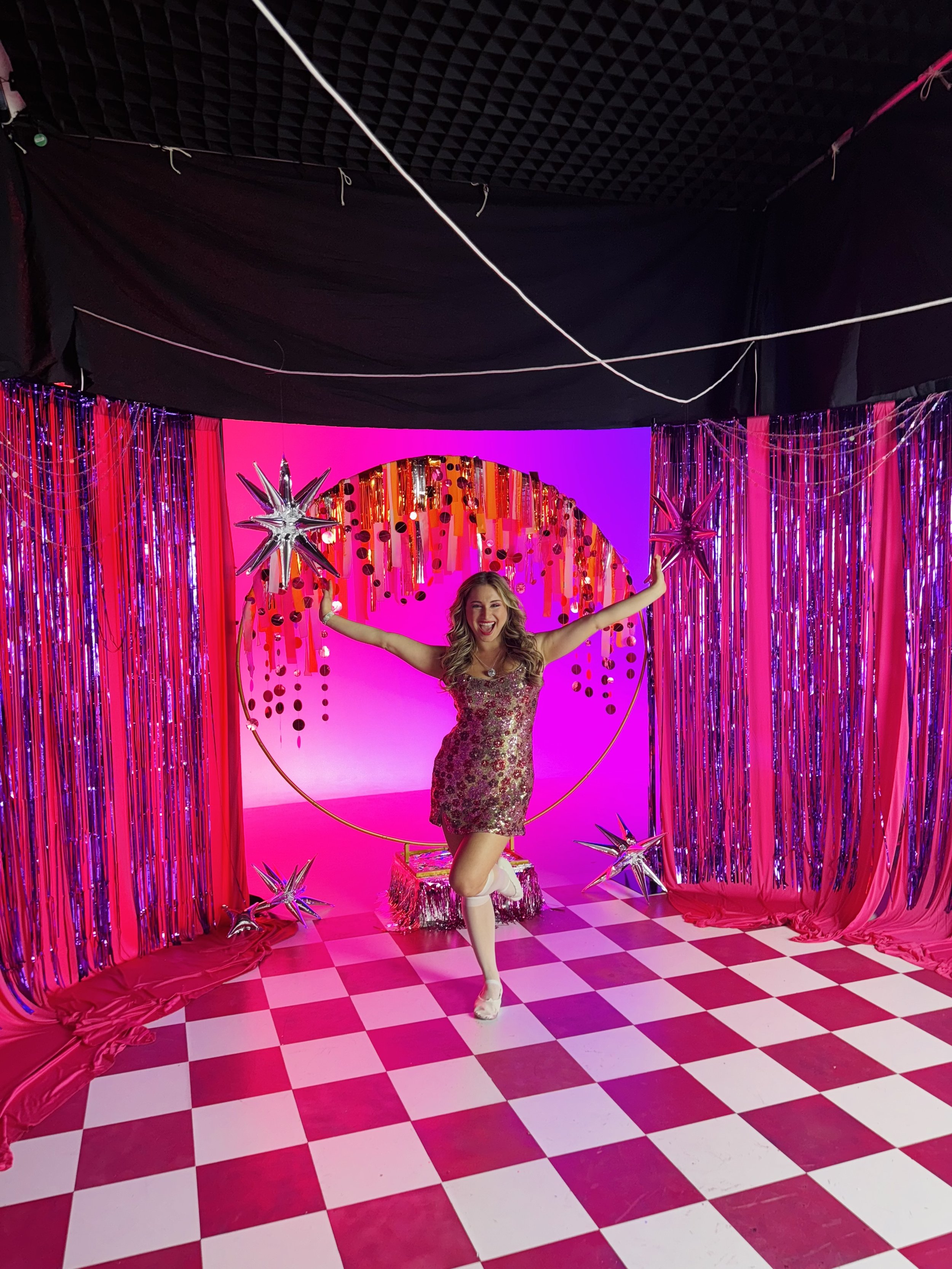 A woman in a sparkly dress celebrating in a colorful, festive setting with pink and purple lighting, silver star balloons, and pink checkered flooring.