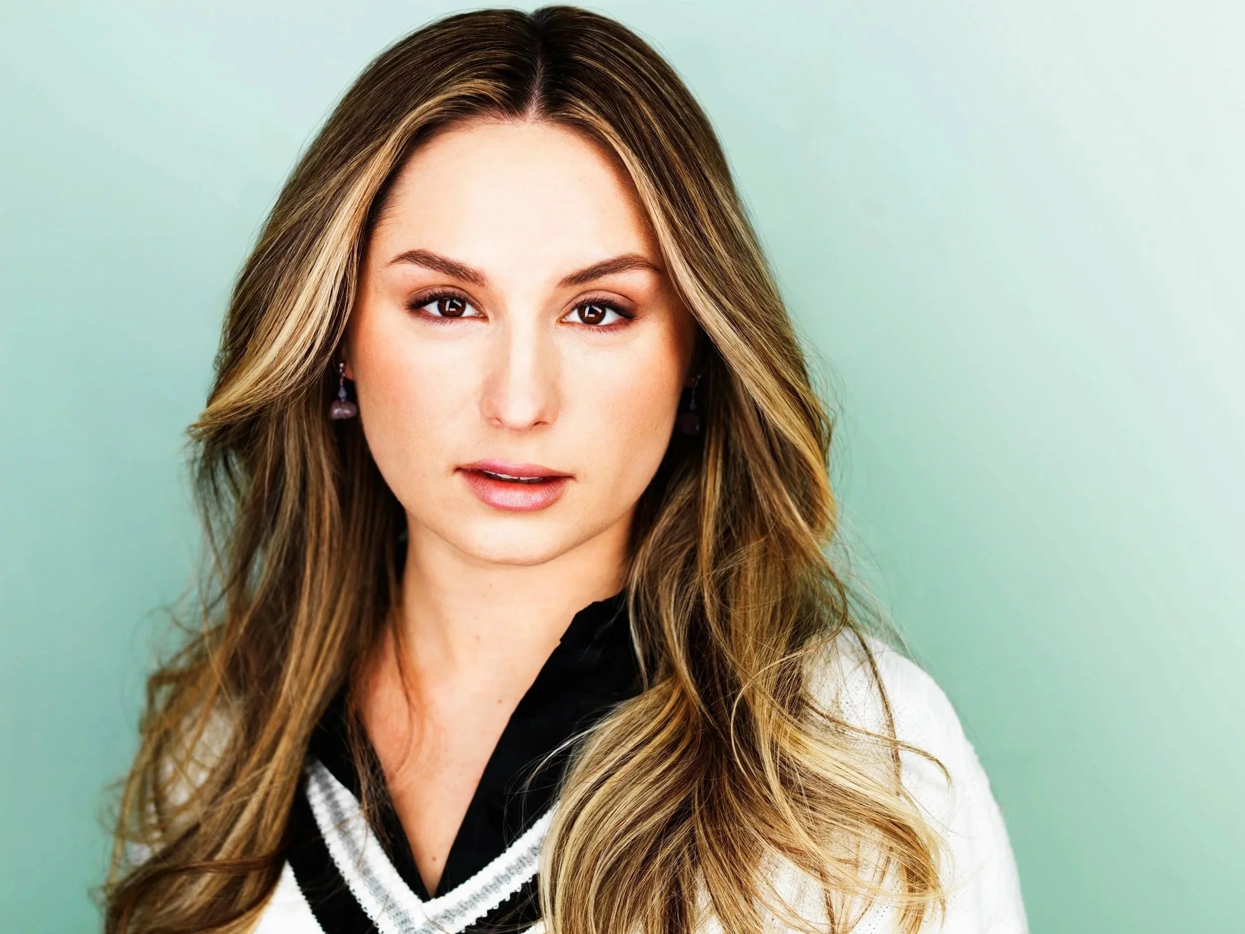 A woman, Karolina Larion, with long, wavy brown hair with blonde highlights, wearing a white sweater over a black  shirt, standing against a light green background.