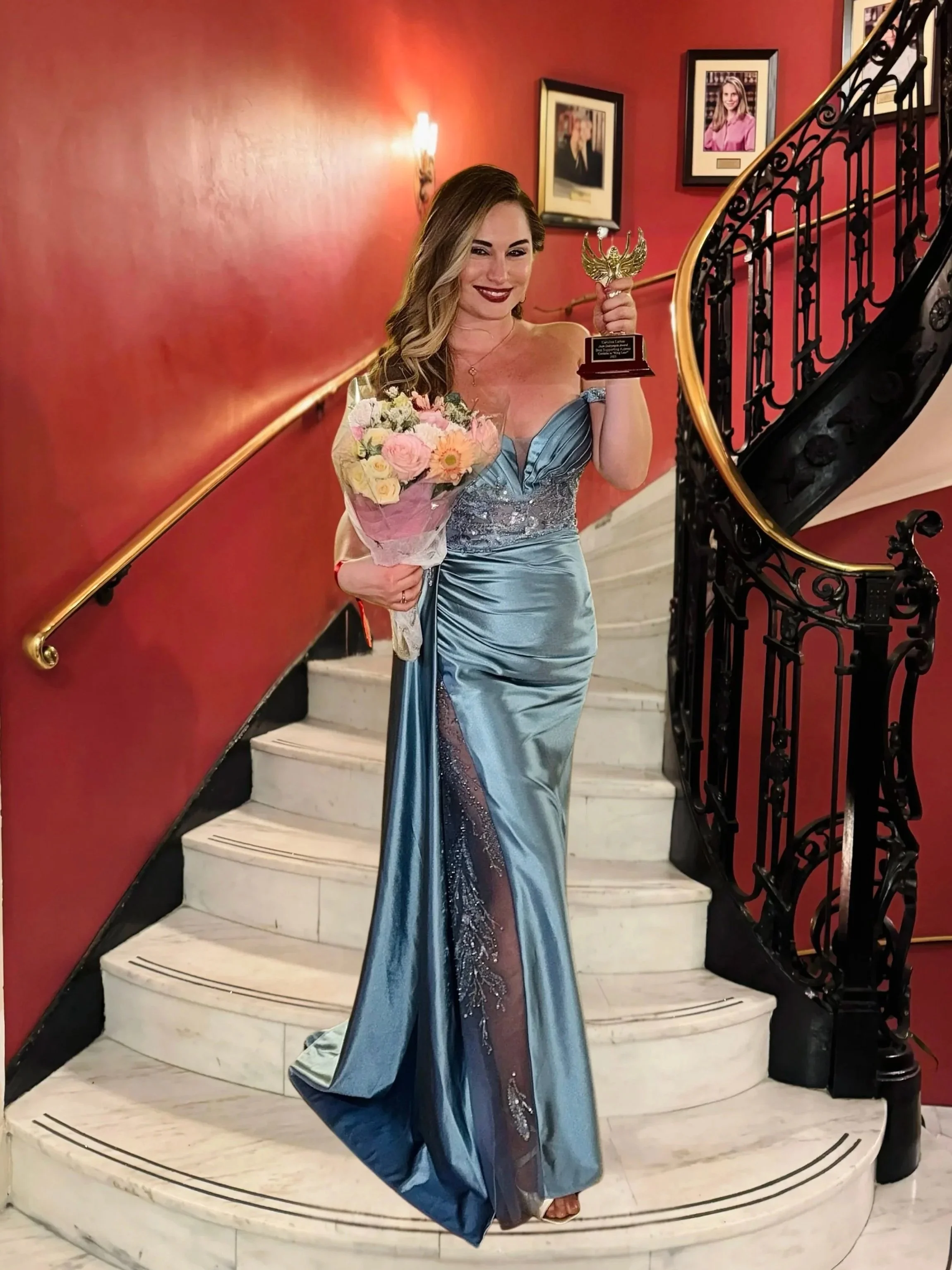 Karolina Larion in a blue evening gown holding a bouquet of flowers and her award for best supporting actress in King Lear, standing on a staircase with framed photos on a red wall behind her.