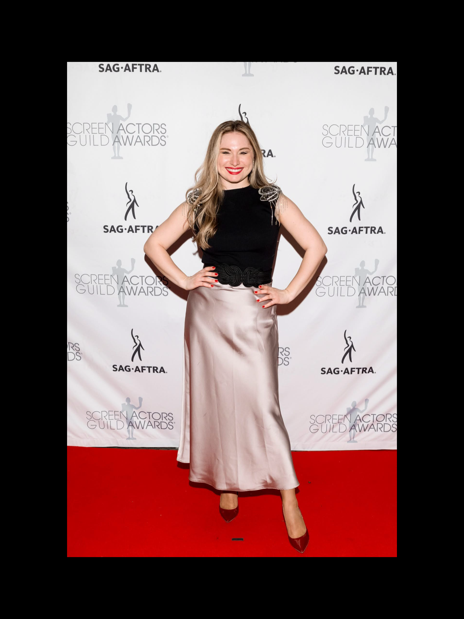 A woman smiling on a red carpet at the Screen Actors Guild Awards, wearing a black sleeveless top with shoulder embellishments, a satin beige skirt, and maroon high heels.