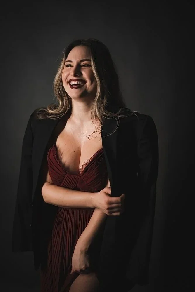 A woman laughing, wearing a red dress and a black blazer, standing against a dark background.