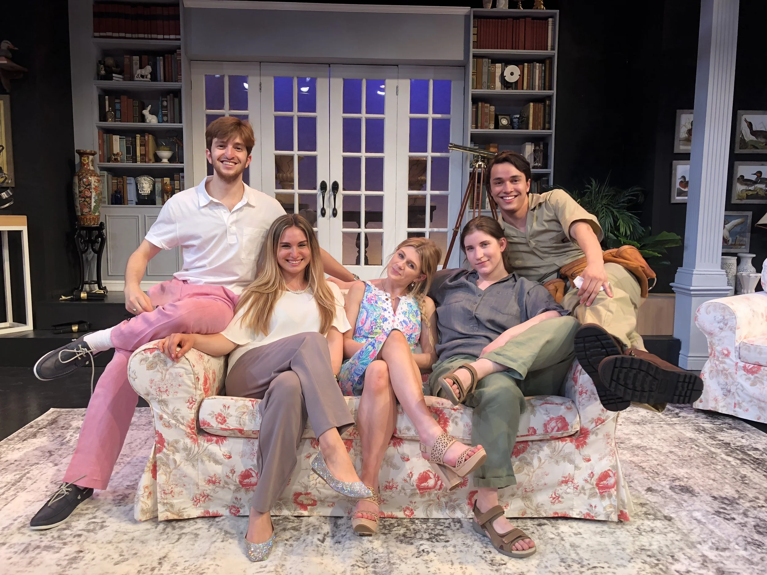 Five young adults sitting and standing on a floral-patterned sofa in a cozy living room, smiling at the camera. The room has bookshelves, decorative vases, artwork, and a telescope in the background.