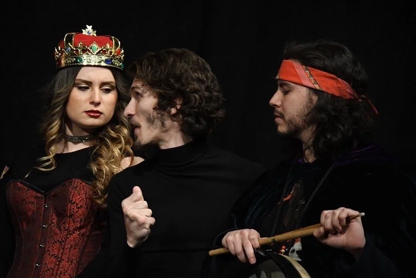 Three people dressed in costumes, one wearing a crown, another in a black turtleneck, and the third with a red bandana, appear to be participating in a theatrical performance or themed photoshoot.