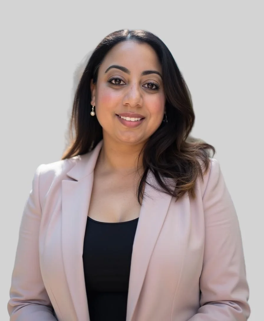 A woman with dark brown hair, brown eyes, and light makeup smiling, wearing pearl earrings, a black top, and a light pink blazer against a plain light-colored background.