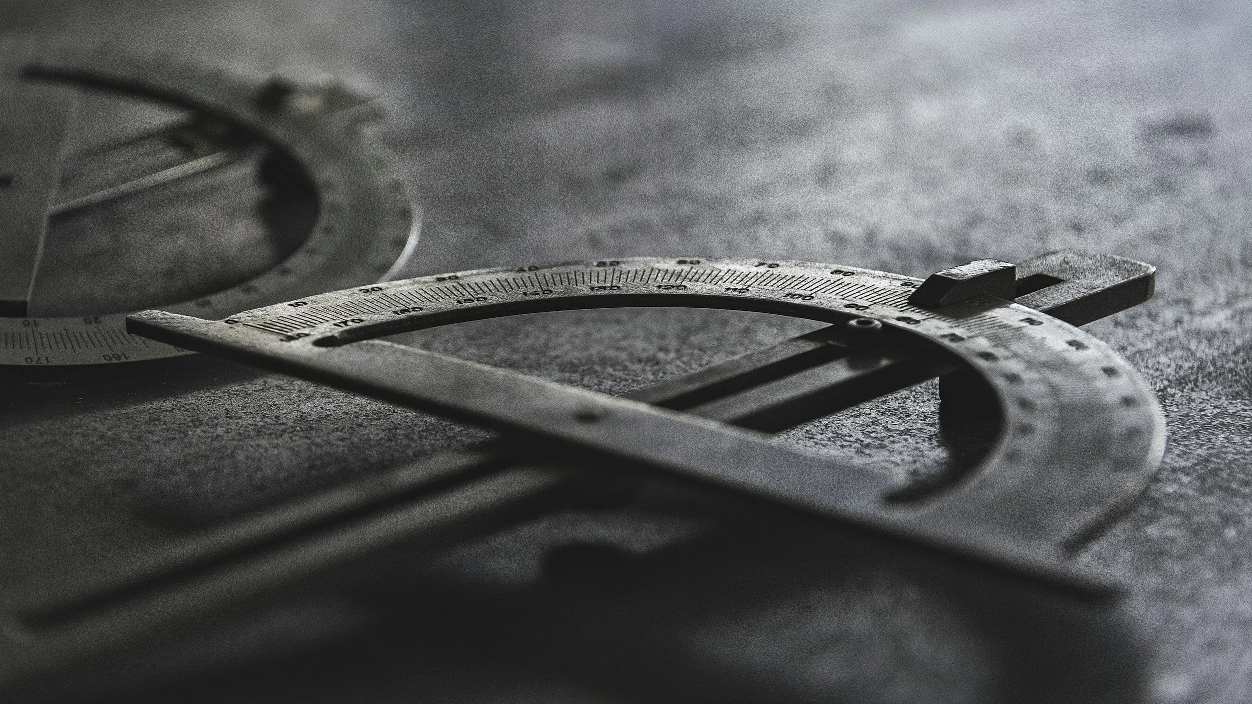 Close-up of a metallic engineering compass lying on a textured gray surface, with part of a protractor in the background to portry accuracy and quality of work.