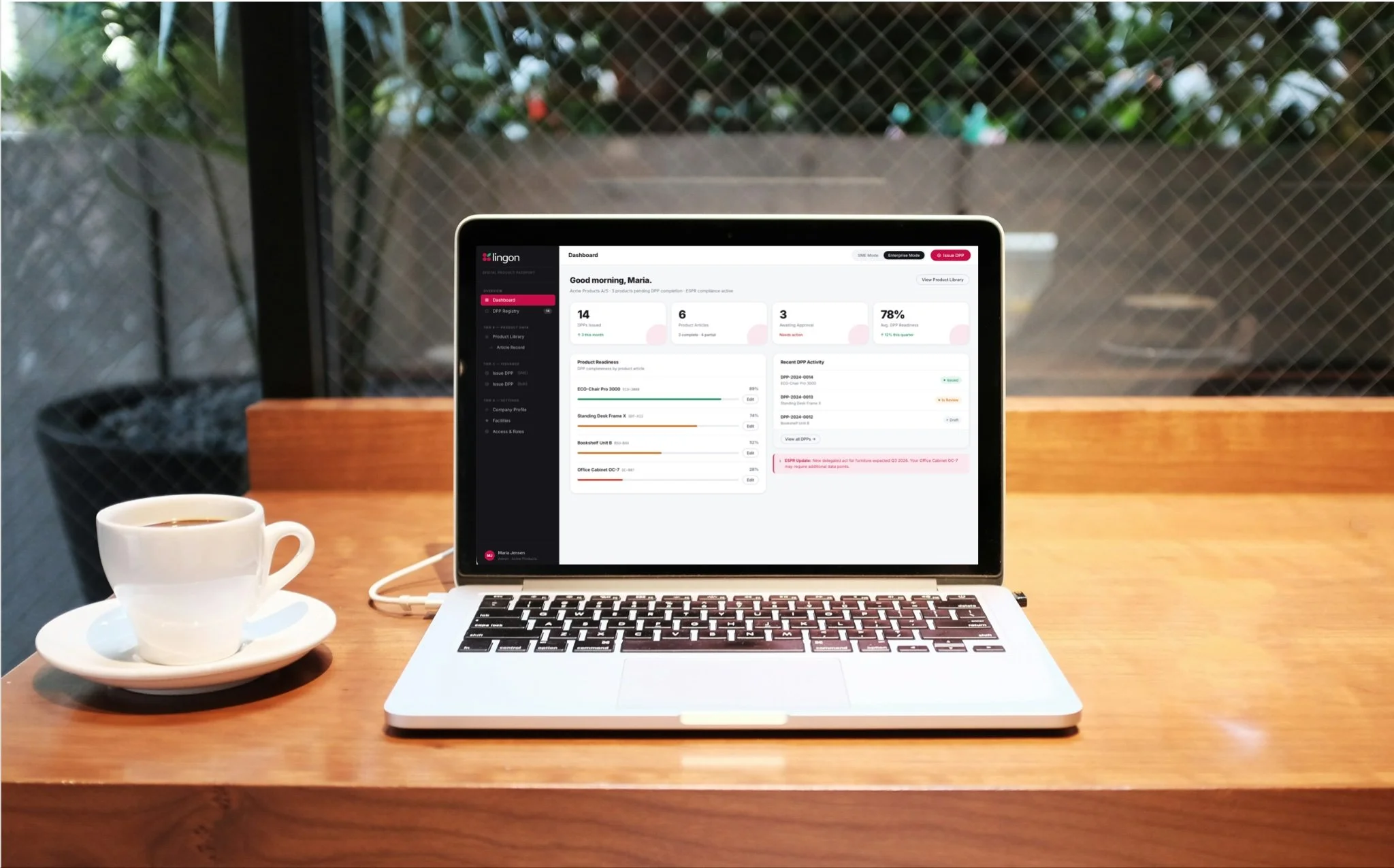 A laptop on a wooden table displaying a dashboard, next to a cup of coffee or tea on a saucer, in front of a large window with a wire mesh or grid in the background.