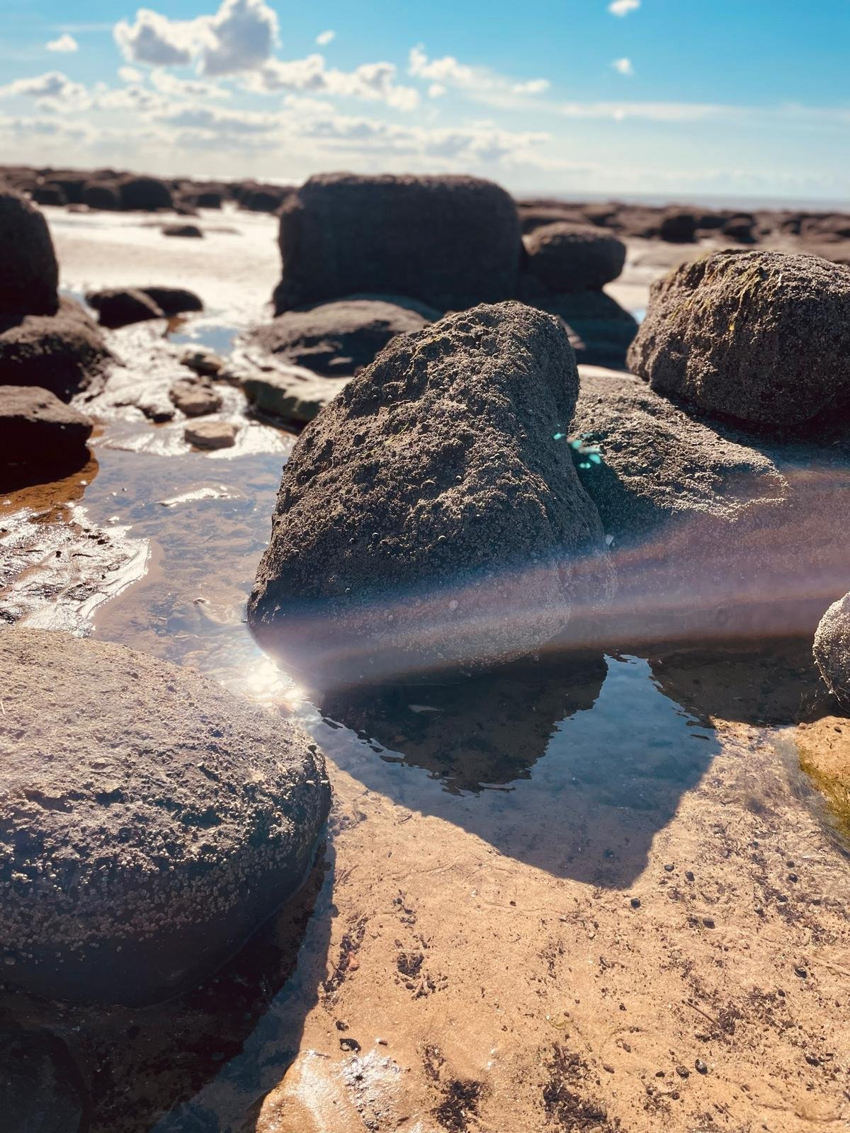 Rocks on a sandy beach with small pools of water, under a partly cloudy blue sky.