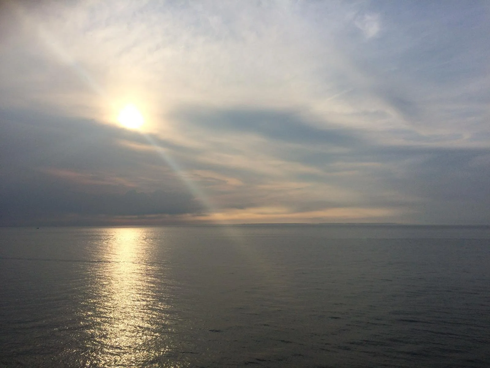 A serene ocean scene with the sun partially obscured by clouds, casting a reflection on the calm water below.
